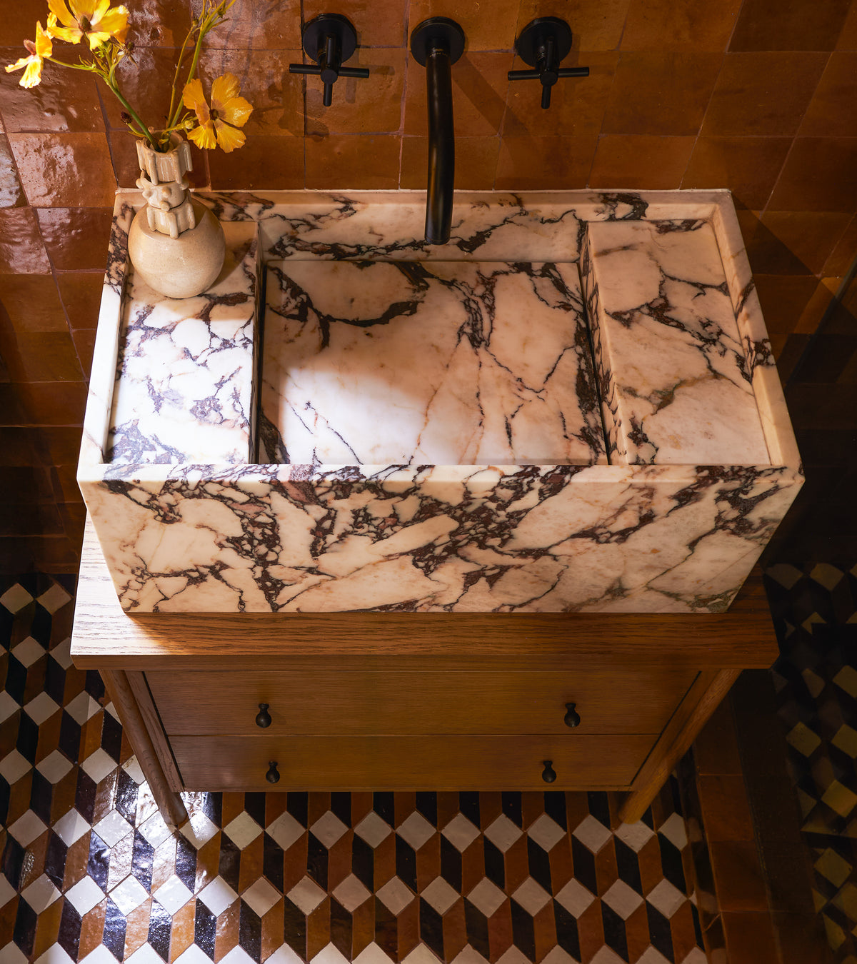 A marble sink with black veining sits on a wooden vanity beneath black wall-mounted faucets. A small vase with yellow flowers is on the left. The floor features Zia Tile’s Prismatic 6, displaying a Moroccan-inspired geometric pattern.
