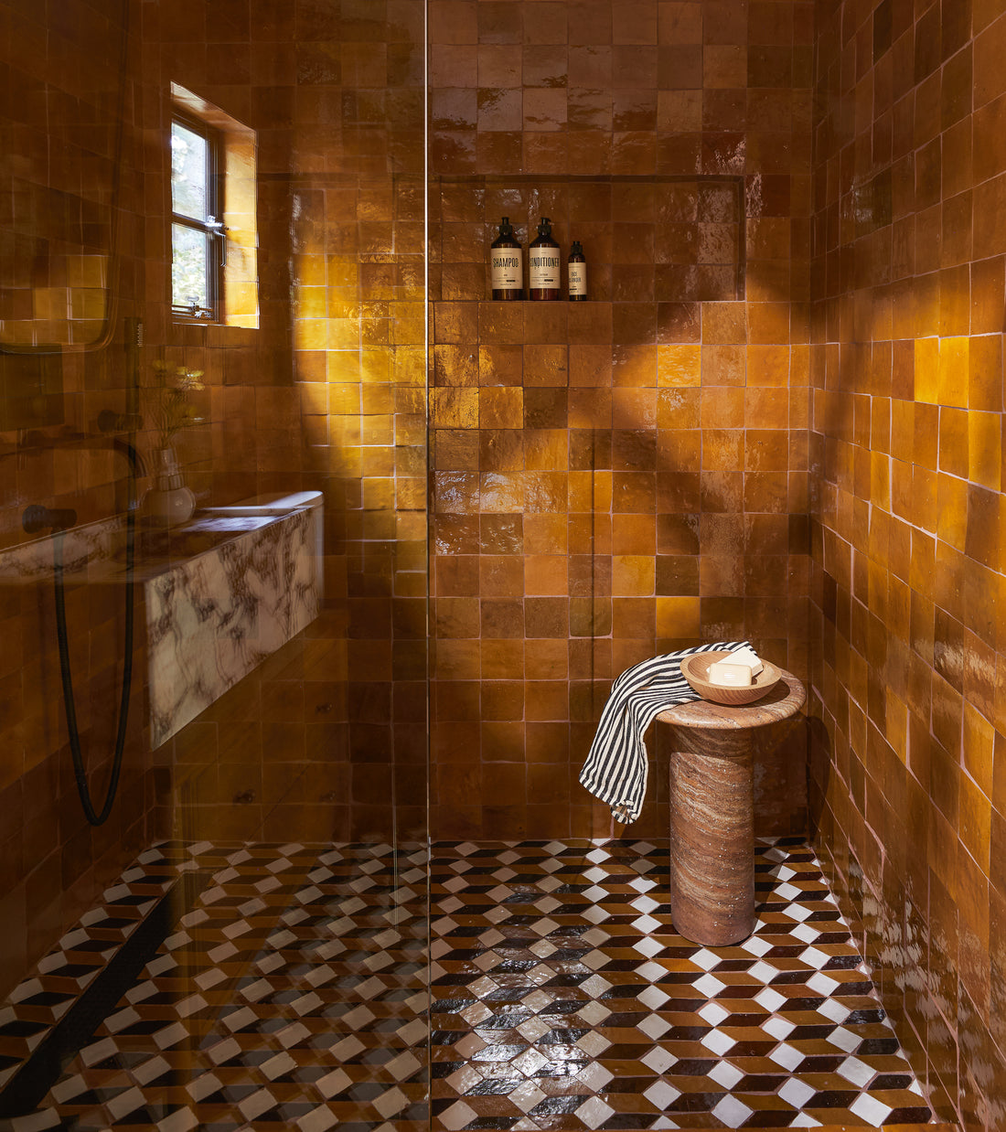 A shower features Zia Tile's Prismatic 6 glossy brown square tiles, a checkered floor, marble shelf and round stool with a striped towel and bowl, plus a niche for bottles. Sunlight streams in through the window.