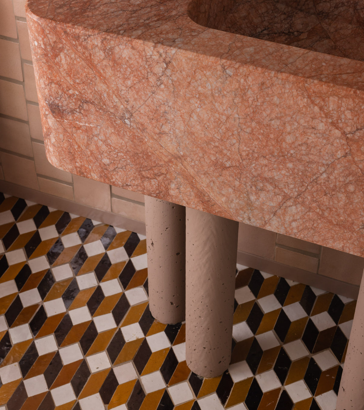 Close-up of a pink marble sink with cylindrical beige legs, set against a wall featuring Zia Tile’s Prismatic 6 mosaic zellige tiles and a geometric floor with brown, yellow, and white 3D cube patterns.