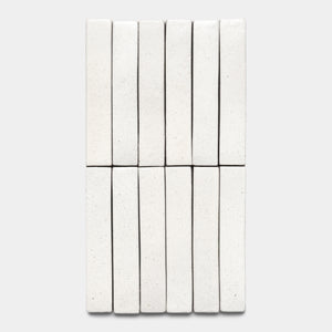 Twelve Puebla Mini Bar tiles by Zia Tile, rectangular and white with speckles, are neatly arranged in a grid on a plain light gray background.