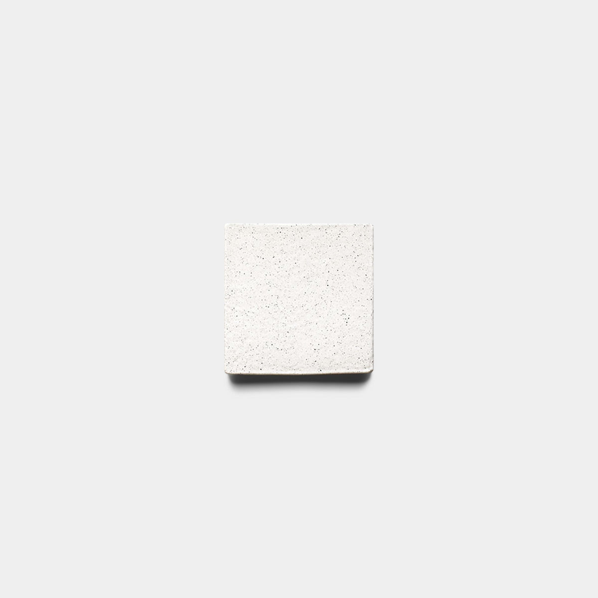 A Zia Tile Puebla 4x4 Square sample, handmade with a smooth speckled white surface and small dark flecks, is centered on a plain light gray background, casting a subtle shadow beneath it.