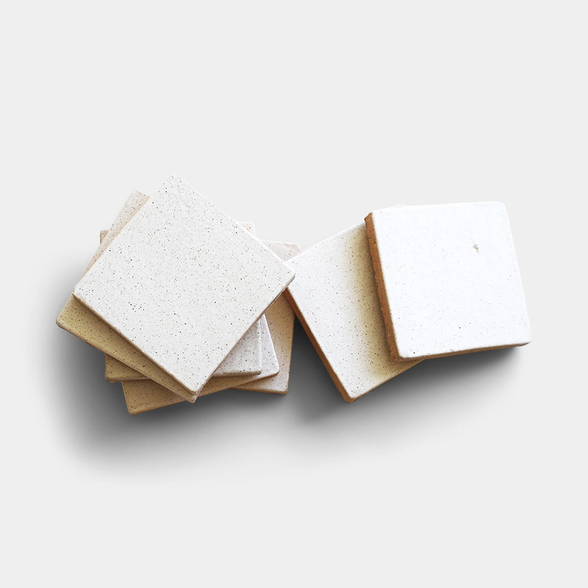 Several Puebla 4x4 Square tiles from Zia Tile—with a square shape, off-white tone, and subtle speckles—are stacked and slightly scattered on a light, neutral background.