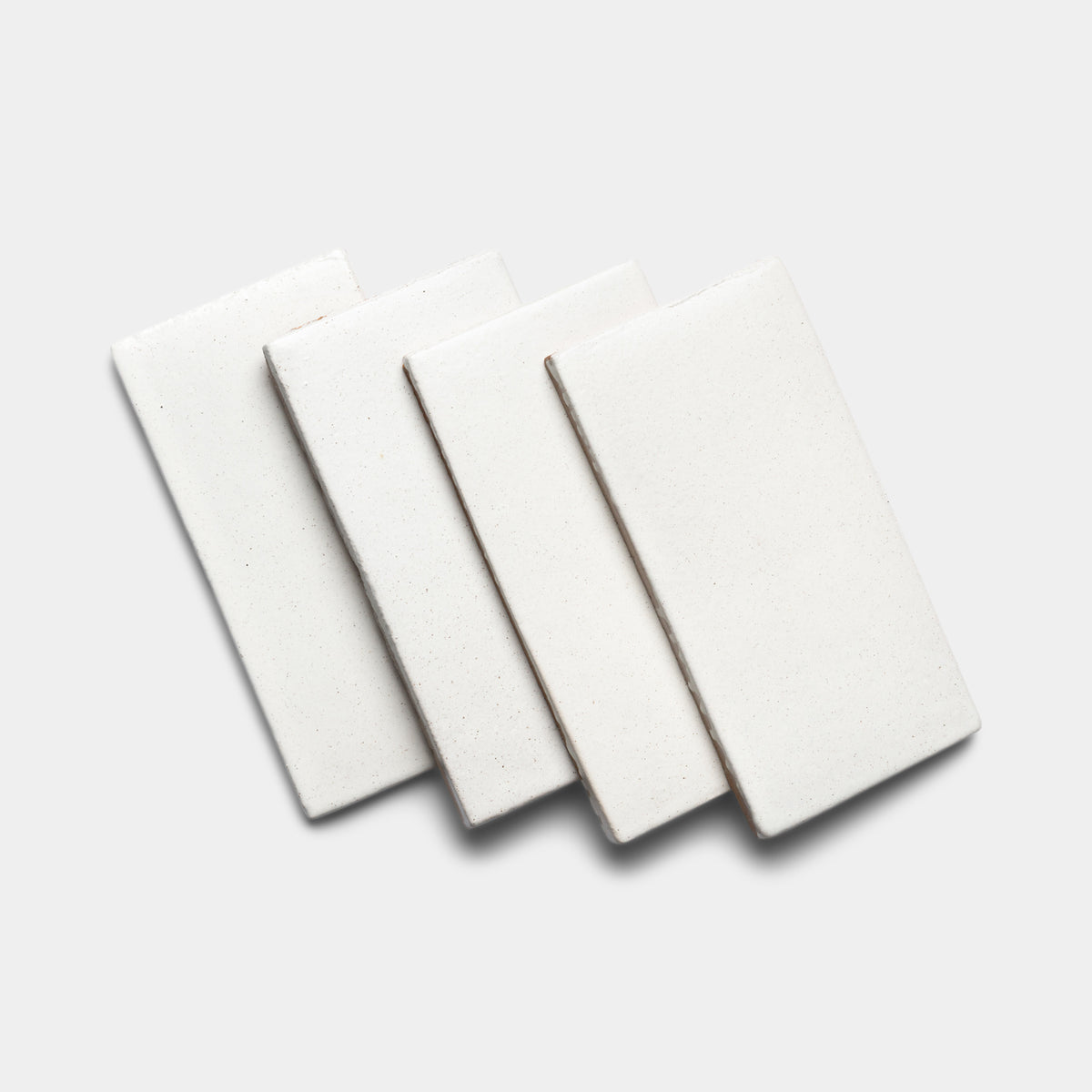 Four white rectangular ceramic tiles are arranged in an overlapping, slightly fanned pattern on a plain light gray background, highlighting the clean style of Zia Tile's Puebla 4x8 Rectangle.