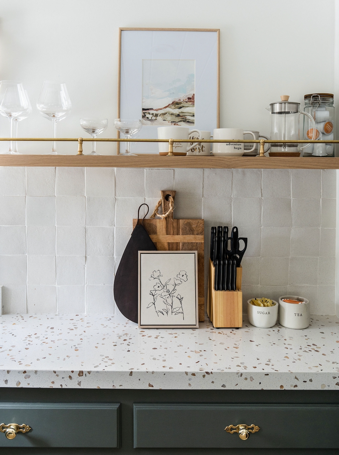 A modern kitchen countertop with cutting boards, a knife block, labeled sugar and tea containers, Cotto jars and mugs sits beneath a shelf against a white Puebla 4x4 tiled backsplash by Zia Tile.