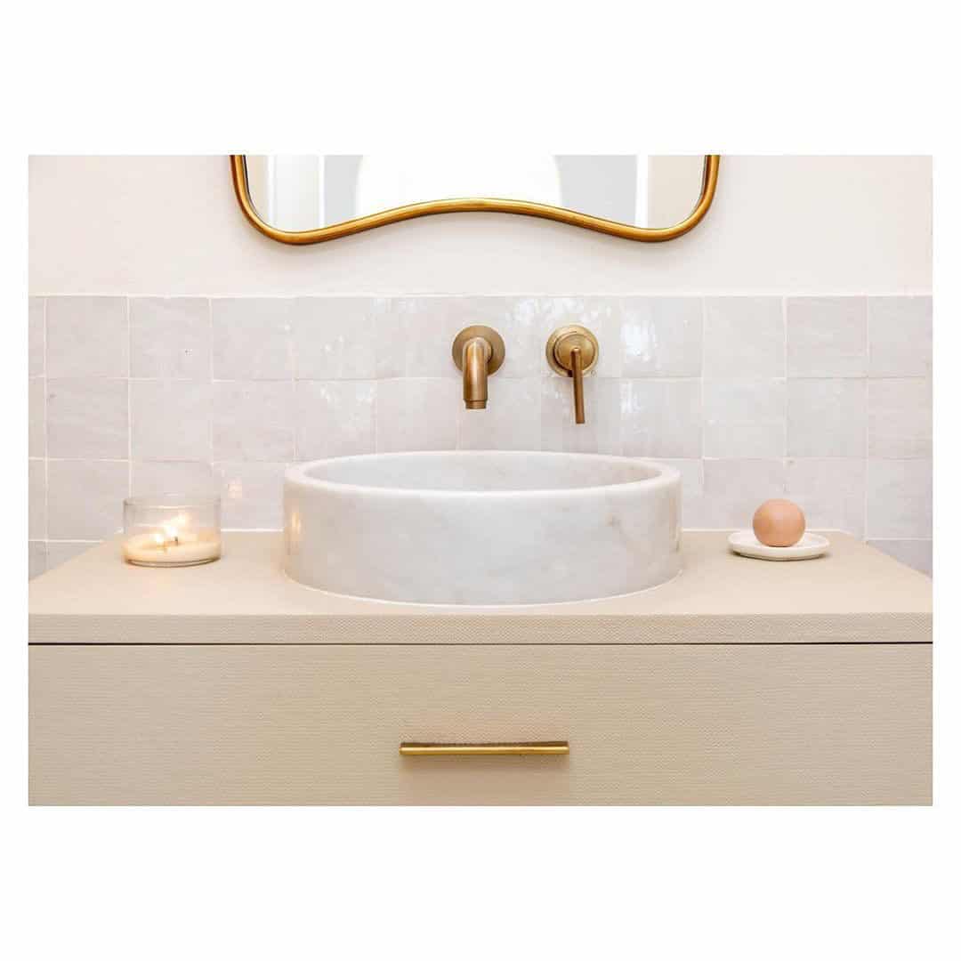 A modern bathroom sink with a round white basin, gold faucet fixtures, Zia Tile Pure White 4x4 tiles on the backsplash, a lit candle on the left, a soap dish with round soap on the right, and a partial gold-framed mirror above.