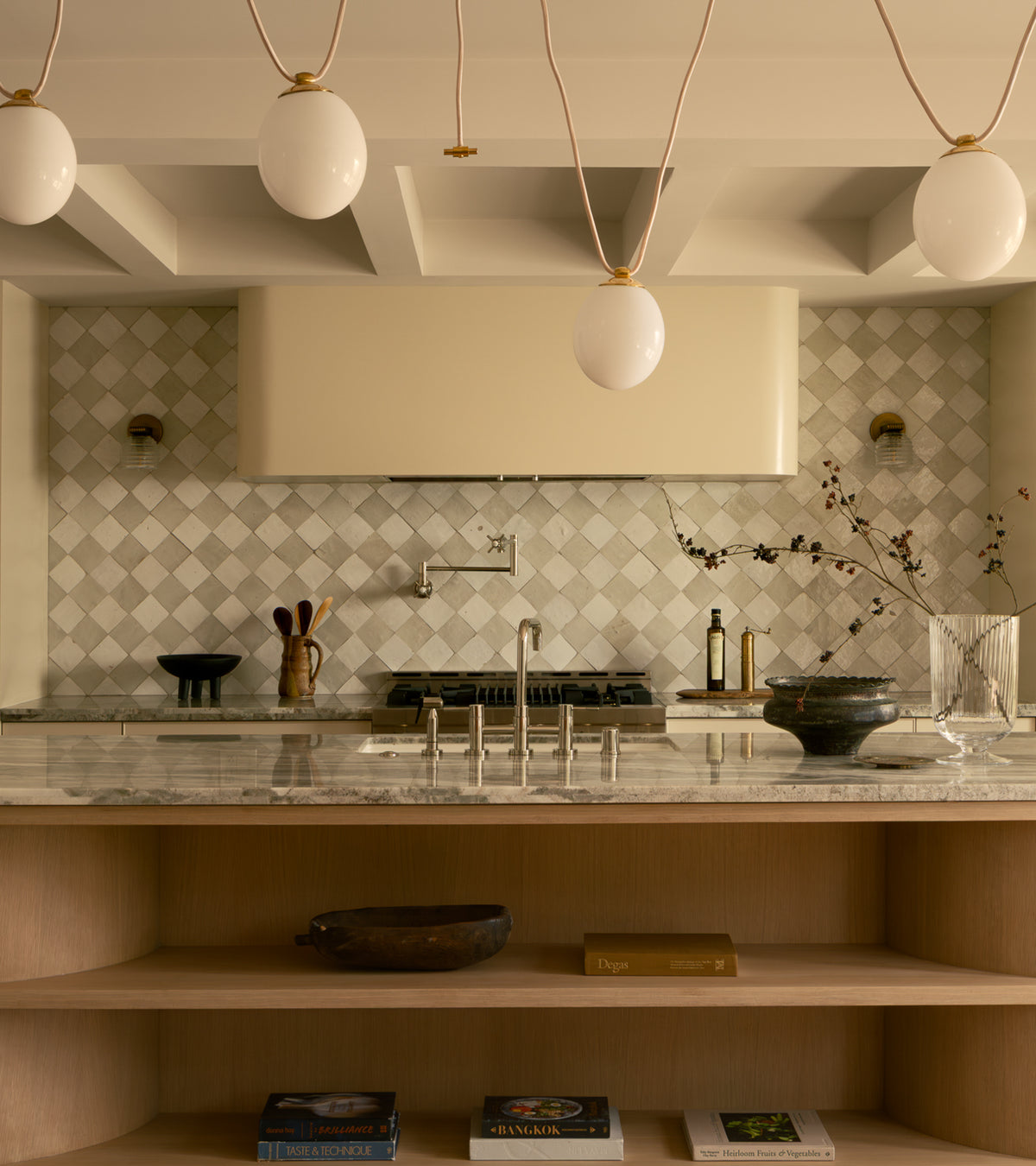 A modern kitchen features a marble island, open wooden shelves with books and bowls, and a stove framed by Zia Tile’s Pure White 4x4 tiles. Four oval pendant lights hang above, while minimalist decor and Moroccan accents complete the look.