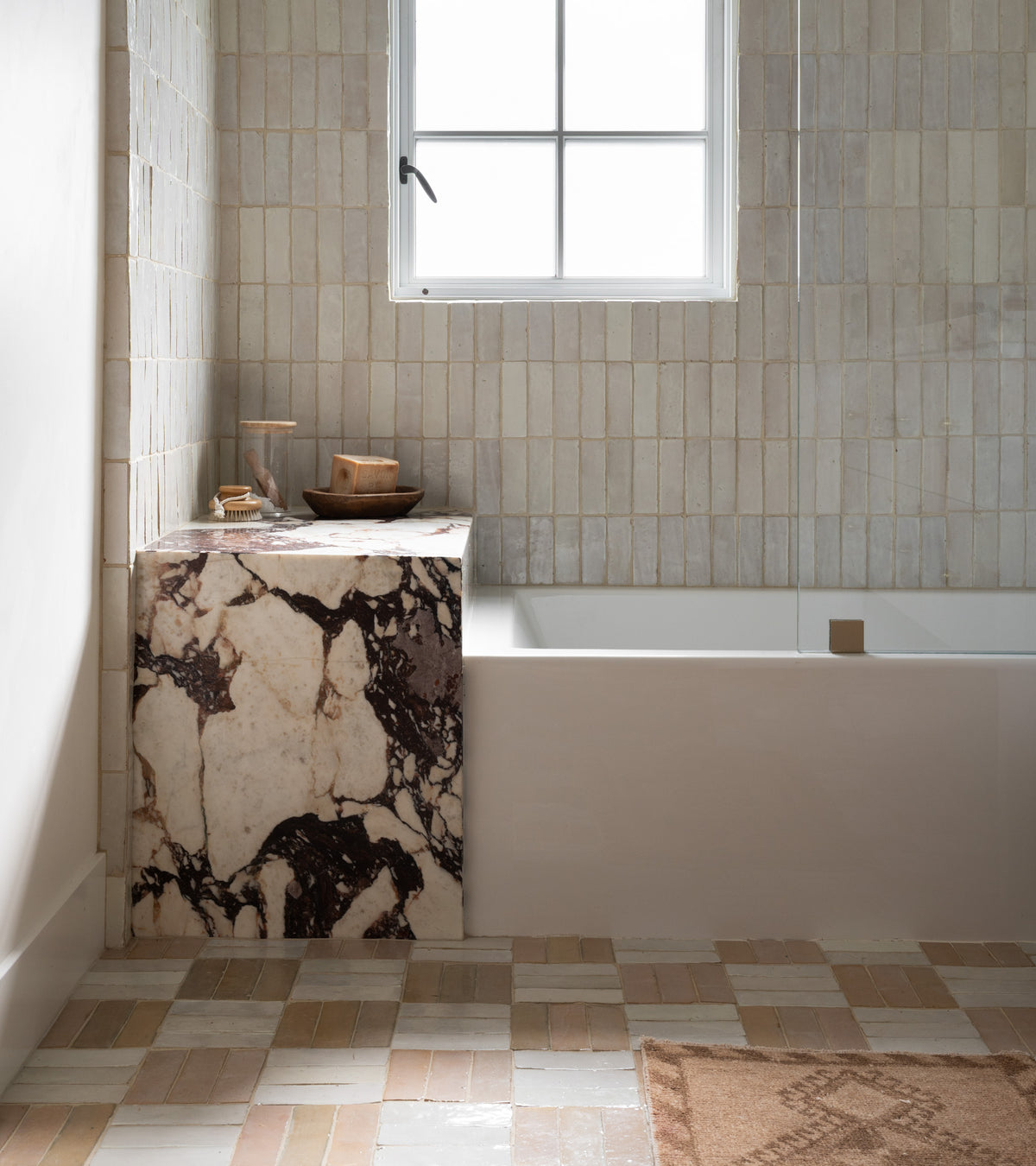 A minimalist bathroom features Zia Tile’s Pure White 2x6 tiles on the walls, a built-in bathtub under a window, marble surround with dark veining, Moroccan-patterned floor tiles, a brown mat, and wood bath accessories.