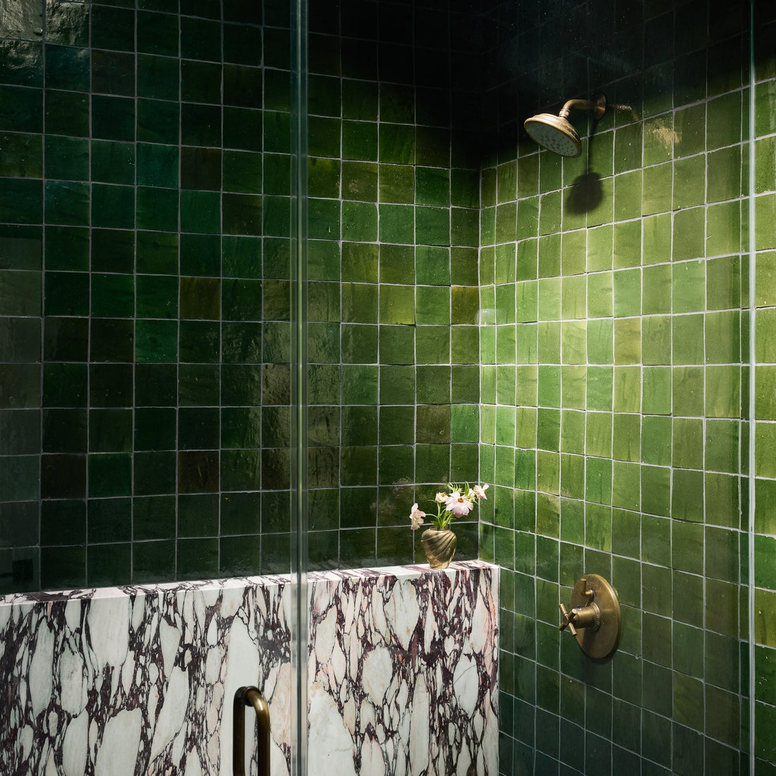 A shower featuring vendor-unknown's Racing Green 4x4 tiles, a brass showerhead and handle, and a white marble partition with dark purple veining. A small vase with flowers rests on the marble ledge.