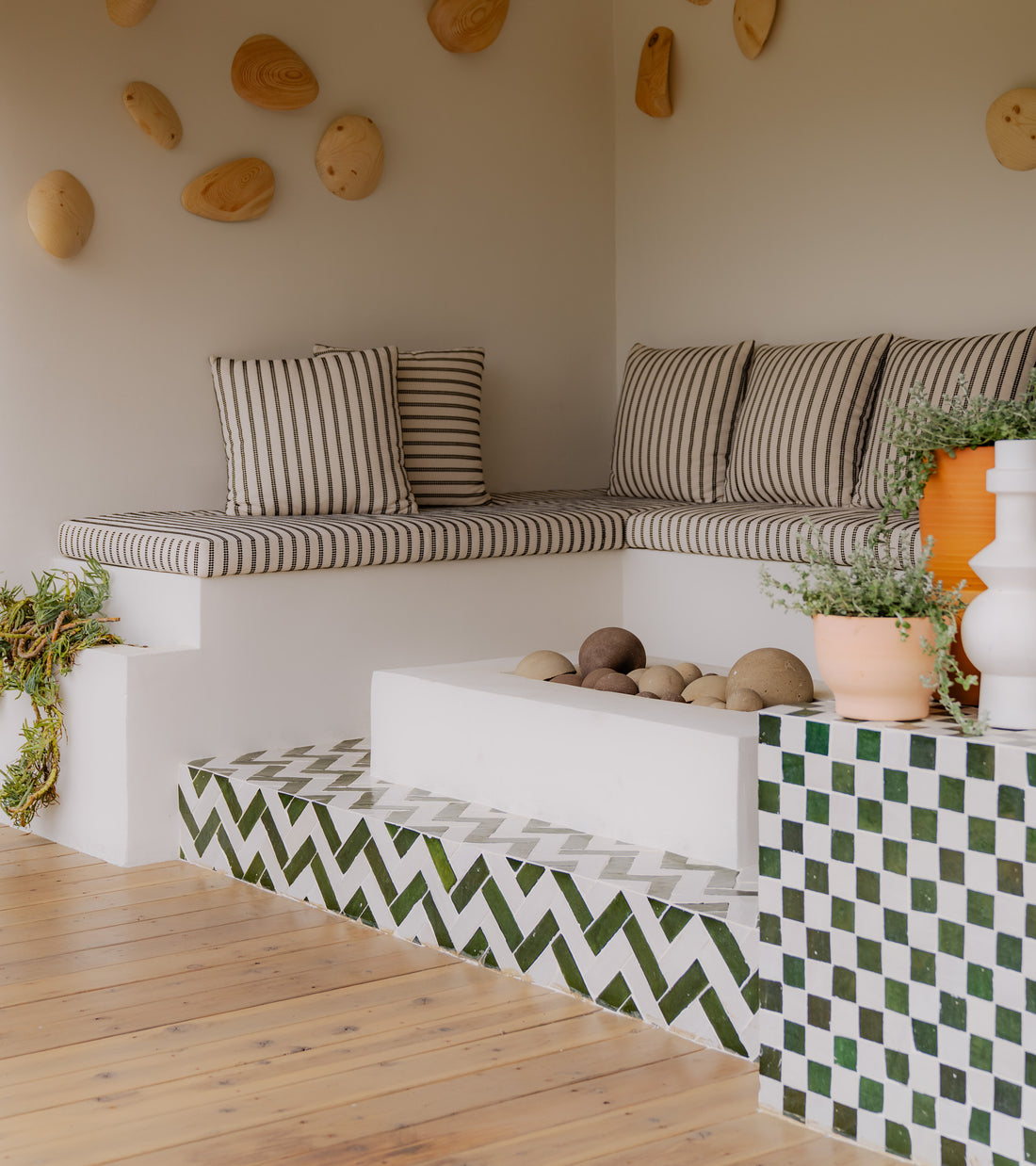 A cozy seating nook with striped cushions, decorative vases, potted plants, and rounded wall accents sits by steps featuring Zia Tile’s Racing Green 2x2 tiles against a light-colored wall.