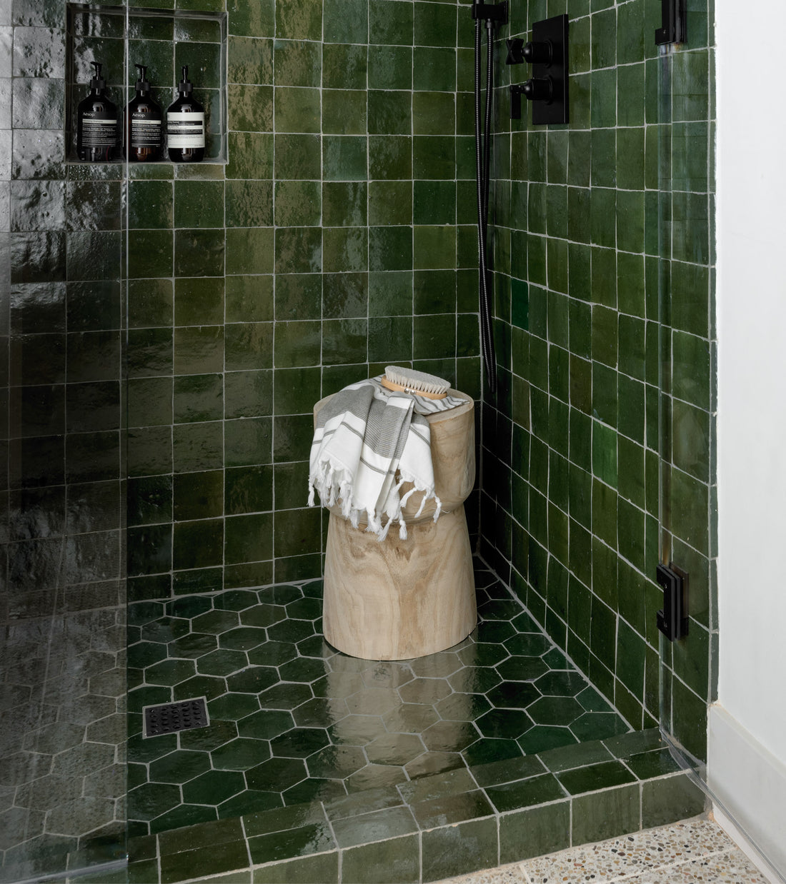 A shower features Zia Tile’s Racing Green Hex zellige tiles and hexagonal floor, with black fixtures, toiletry bottles in a wall niche, plus a small wooden stool holding folded towels and a brush.