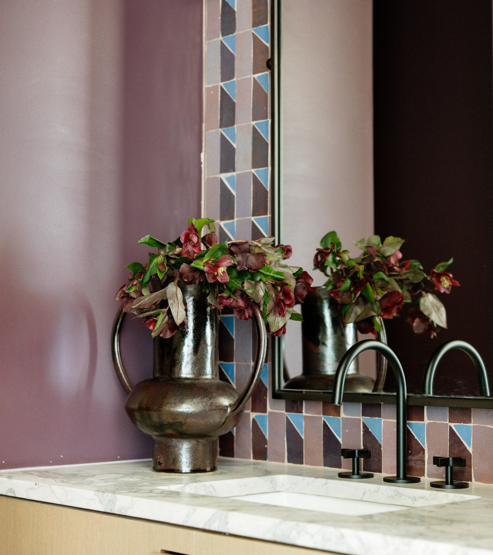 The Radian Inline 3 by Zia Tile features geometric blue and mauve mosaic zellige tiles, creating a vibrant purple-accented wall around the mirror above a marble bathroom counter with a dark bronze vase and black faucet.