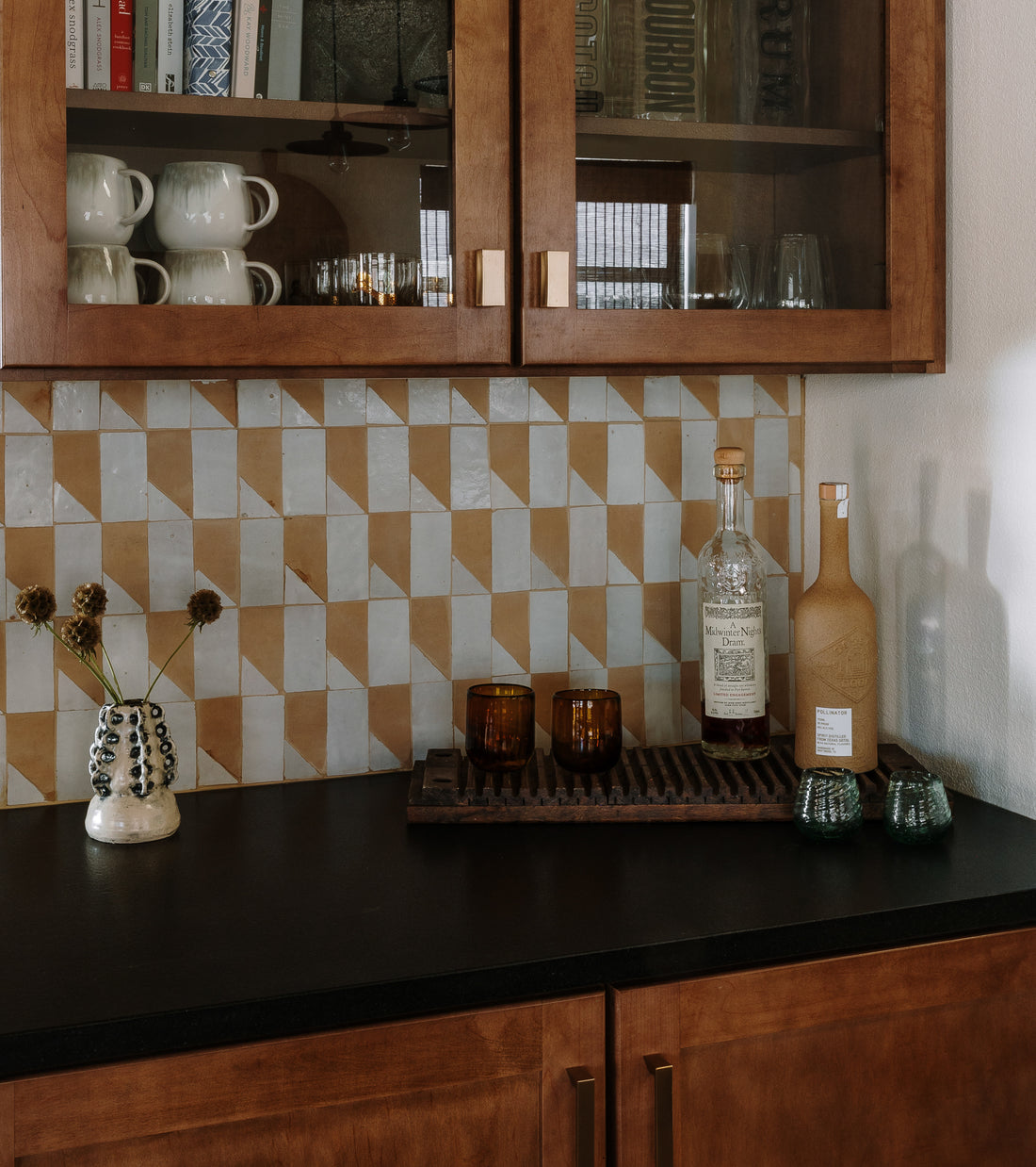 The Radian Offset 1 geometric beige and white Moroccan tile backsplash by Zia Tile adds style above the kitchen counter, which displays bottles, glasses, a tray, and a small vase with dried flowers beneath wooden cabinets.