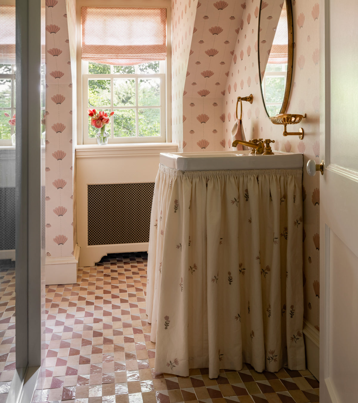 A cozy bathroom features Radian Offset 2 mosaic zellige floor tiles by Zia Tile, pink patterned wallpaper, a round mirror, a skirted sink with gold fixtures, and a window with red flowers. Soft light filters through the blinds.