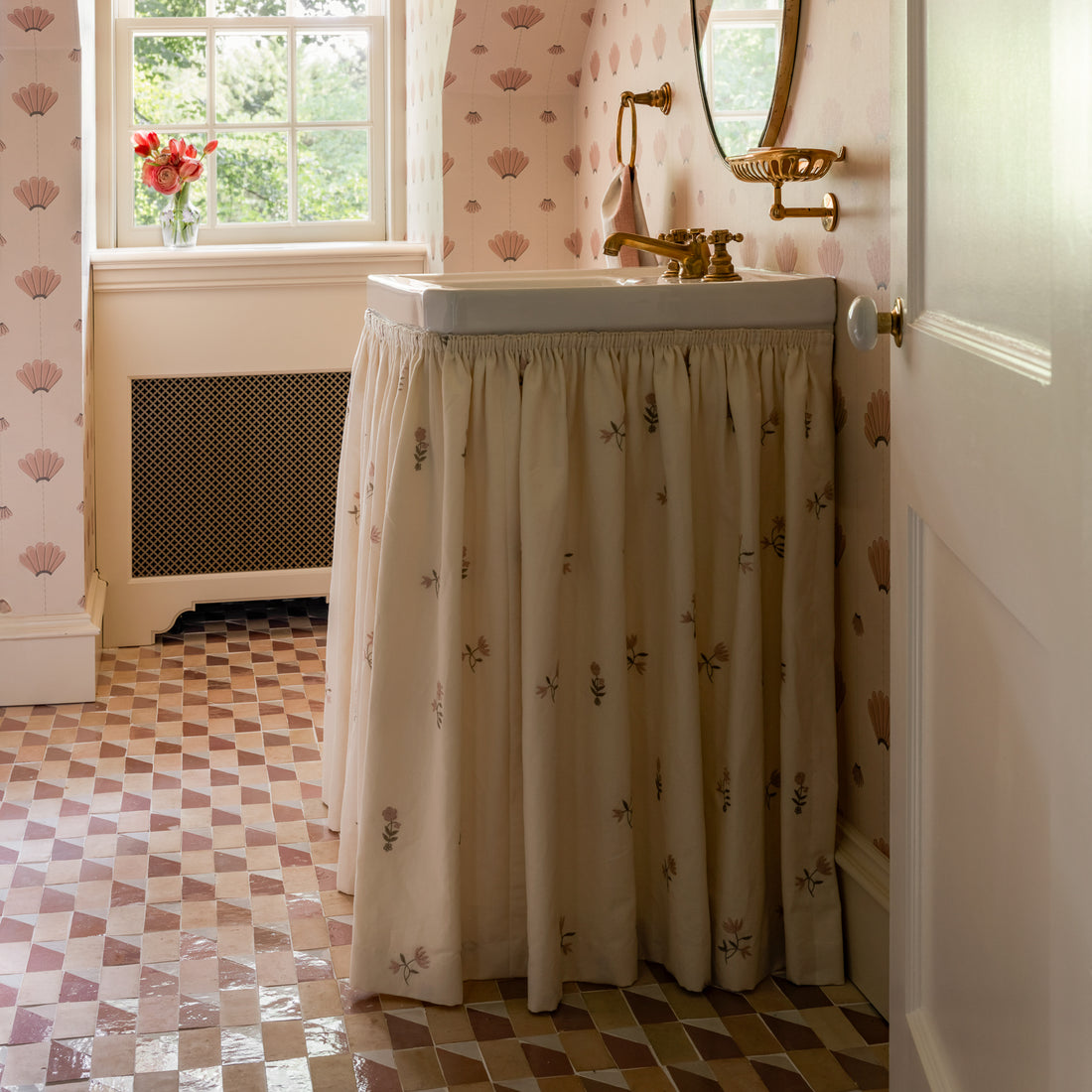 A vintage-style bathroom with a patterned tile floor, a sink with a ruffled fabric skirt, floral wallpaper, a round mirror, and a vase of flowers on a windowsill letting in natural light.