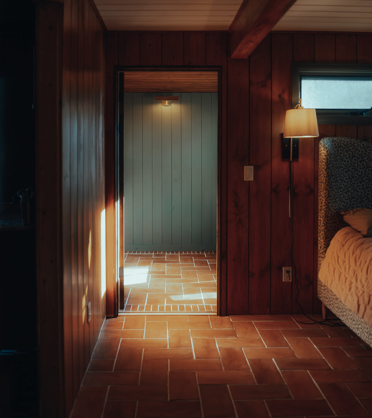 A warmly lit bedroom with wooden walls and 6.5x13 Rectangle + Red Clay tiles by Zia Tile on the floor. Sunlight streams through a doorway, illuminating the handmade tiles. A bed, patterned headboard, and wall lamp are visible on the right.