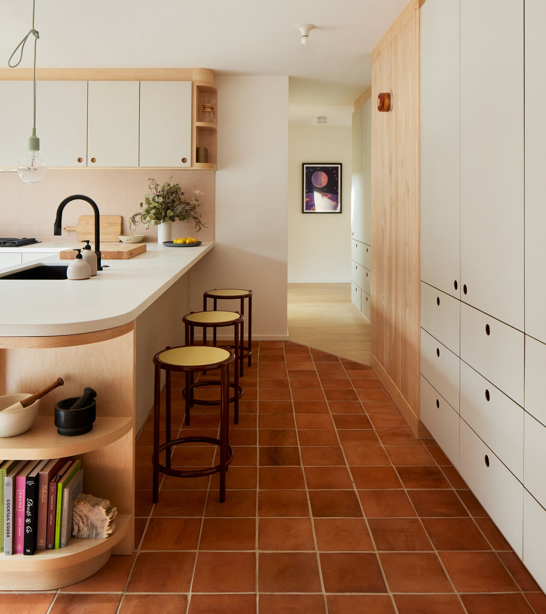 A modern kitchen features 8x8 Square + Red Clay terracotta tile floors by Zia Tile, light wood cabinetry, white countertops with built-in shelves, two round stools, and decor; a hallway with wall art appears in the background.
