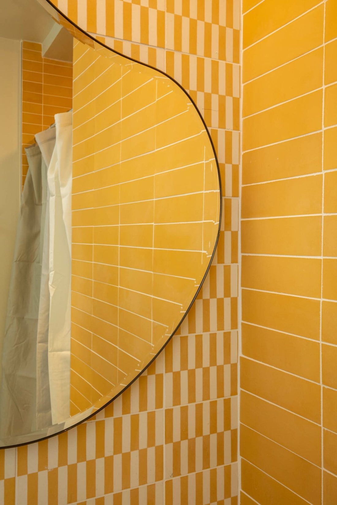 A curvy mirror reflects Zia Tile's Rialto Cadmium 4x8 yellow tiles and part of a white shower curtain in a bathroom with yellow and white checkerboard walls over elegant cement tile flooring.