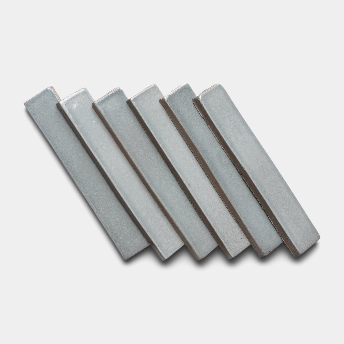 Six Roca Mini Bar tiles by Zia Tile, in light gray ceramic with a rectangular shape, are arranged diagonally in a staggered, overlapping pattern on a plain white background.