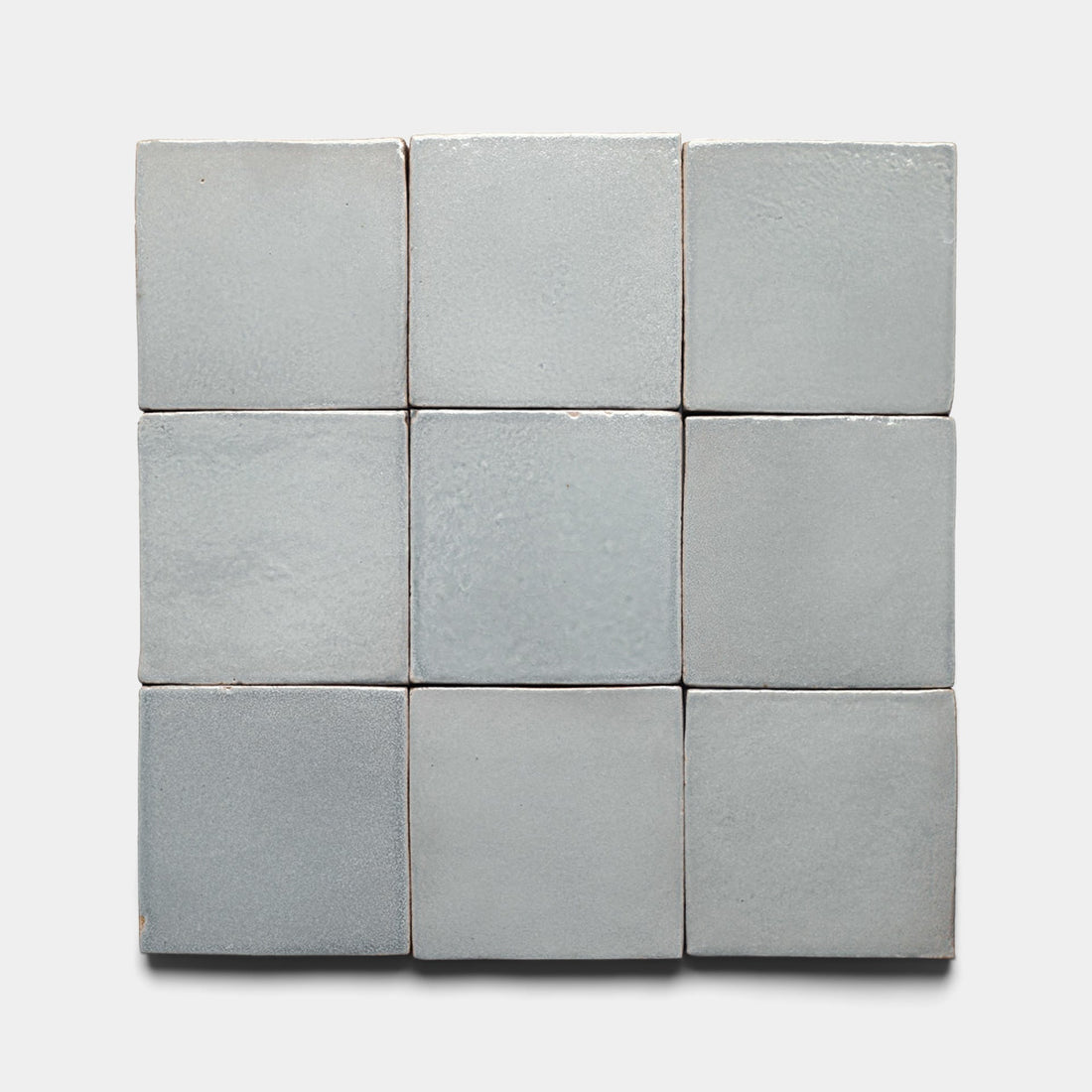 Nine Roca 4x4 Square tiles by Zia Tile, light gray and handmade with a slightly glossy, textured surface and subtle shading variations, are arranged in a 3x3 grid on a plain white background. Inspired by San Miguel de Allende craftsmanship.