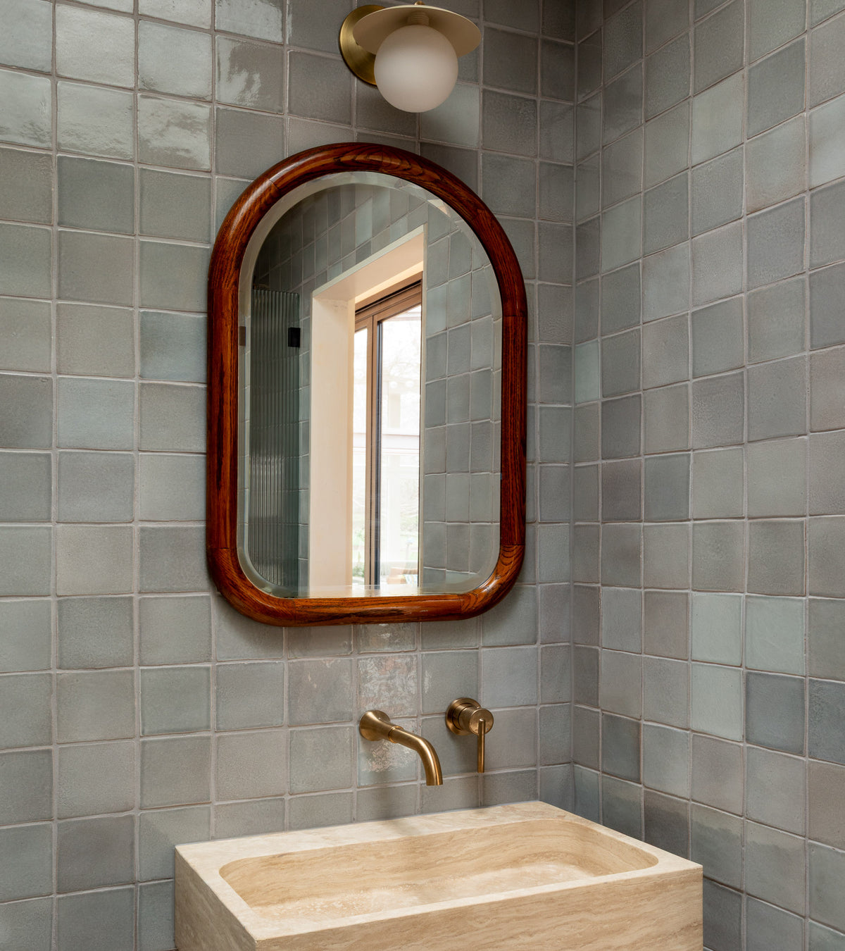 A bathroom features Zia Tile’s Roca 4x4 light gray square tiles, a rectangular beige stone sink, brass wall-mounted faucet, an arched wooden-framed mirror inspired by San Miguel de Allende reflecting a window, and a round white sconce above.