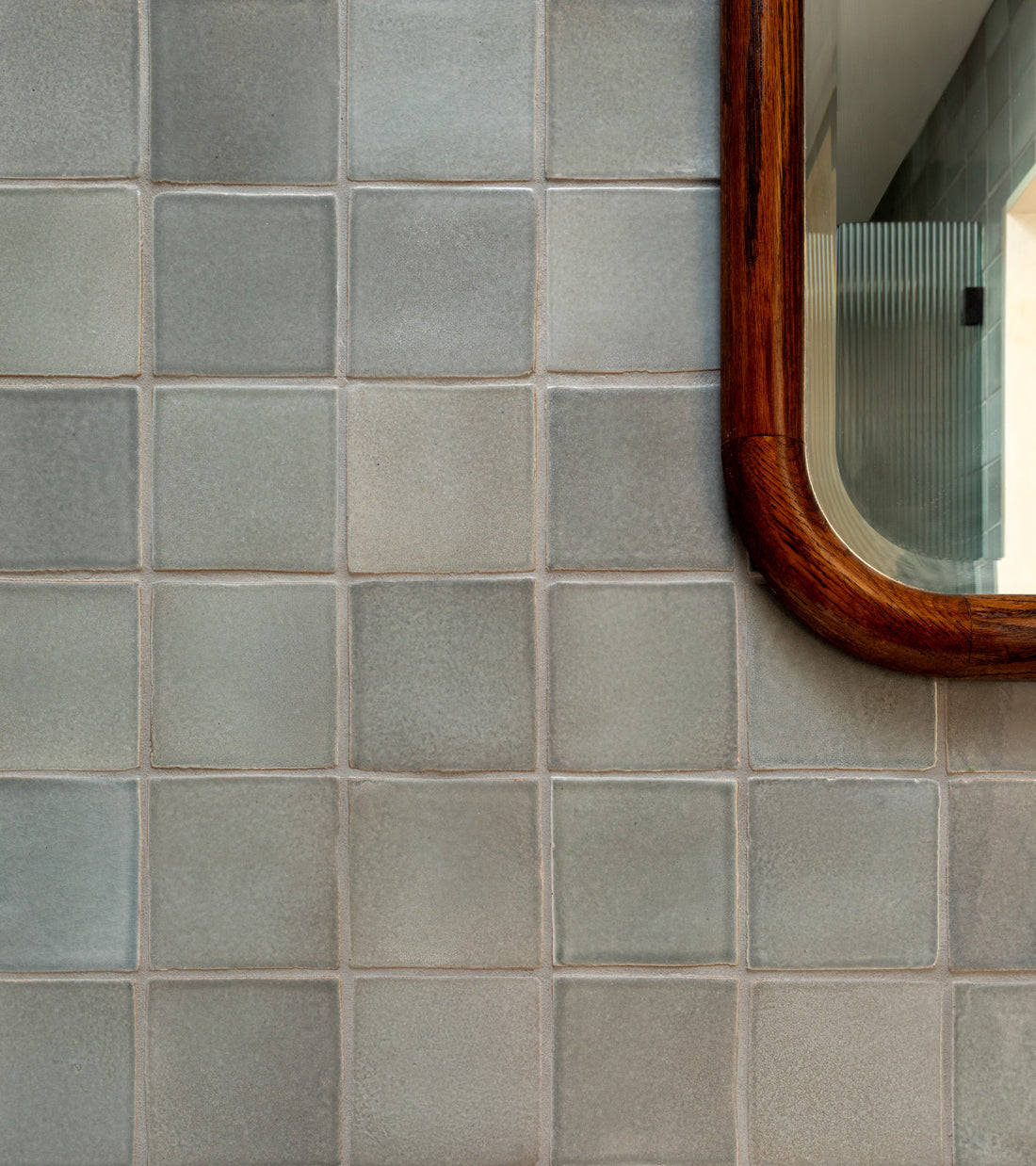 A close-up of a tiled wall featuring Roca 4x4 Square tiles by Zia Tile. On the right, a wooden-framed mirror reflects another handmade Cotto tiled wall and a corridor inspired by San Miguel de Allende.