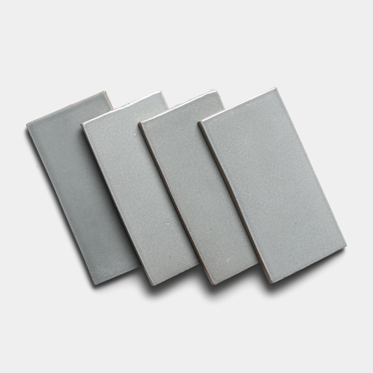 Four Roca 4x8 Rectangle tiles by Zia Tile, featuring a light gray ceramic finish, are arranged in a slightly overlapping diagonal pattern on a plain white background.