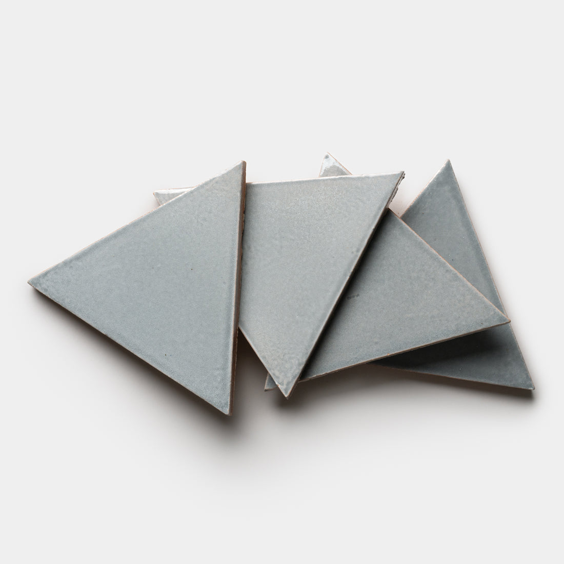 Four Roca Triangle tiles by Zia Tile in gray ceramic are overlapping each other on a white background.