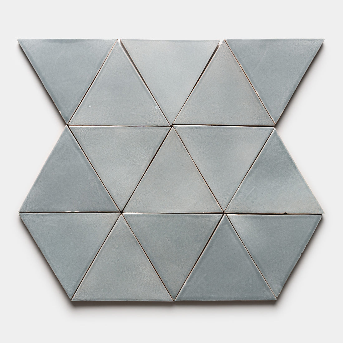 Twelve Roca Triangle tiles by Zia Tile in light gray are arranged on a plain, light gray surface, forming a symmetrical hexagonal pattern.