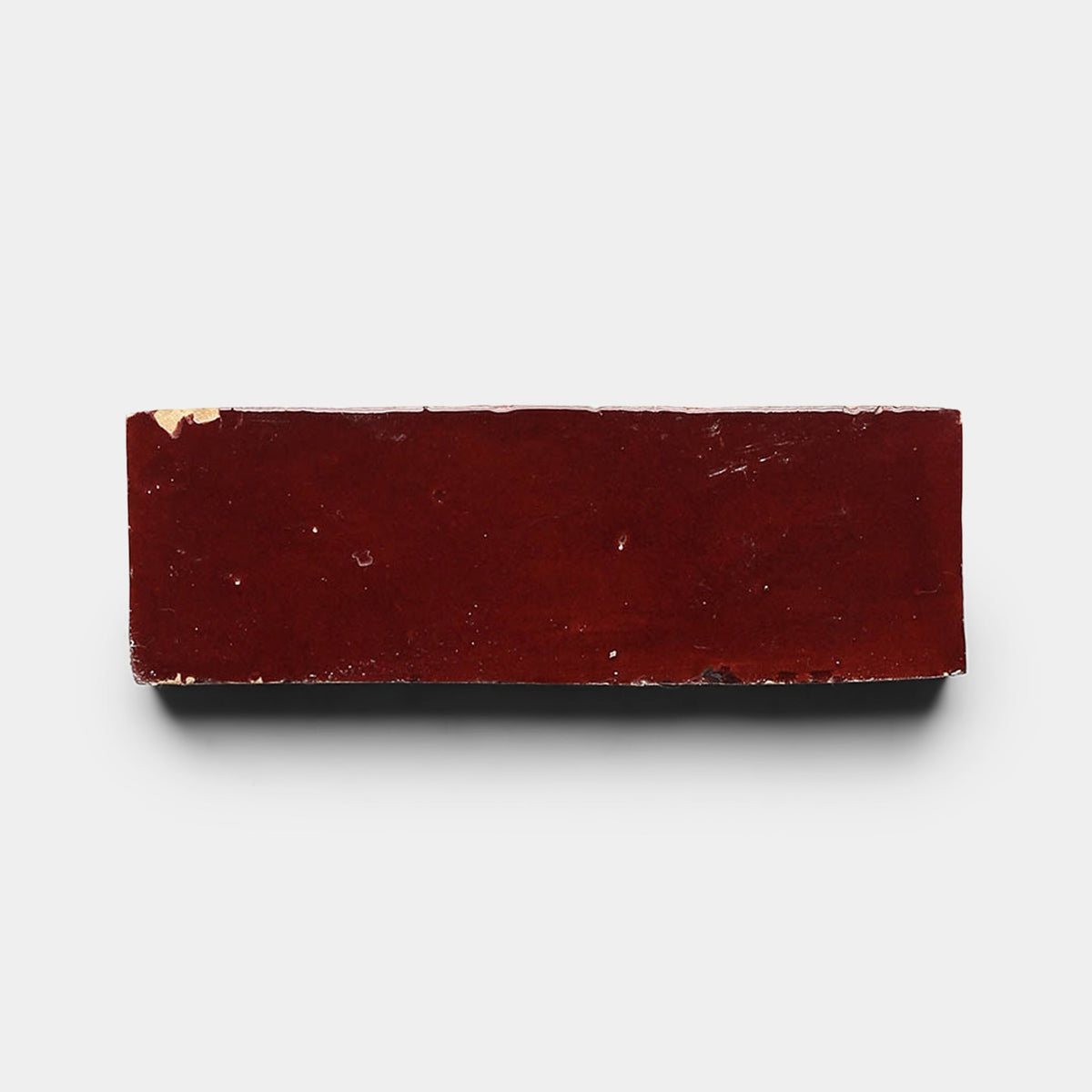 A Rouge 2x6 sample brick by Zia Tile with a chipped corner is photographed on a white background, casting a slight shadow.