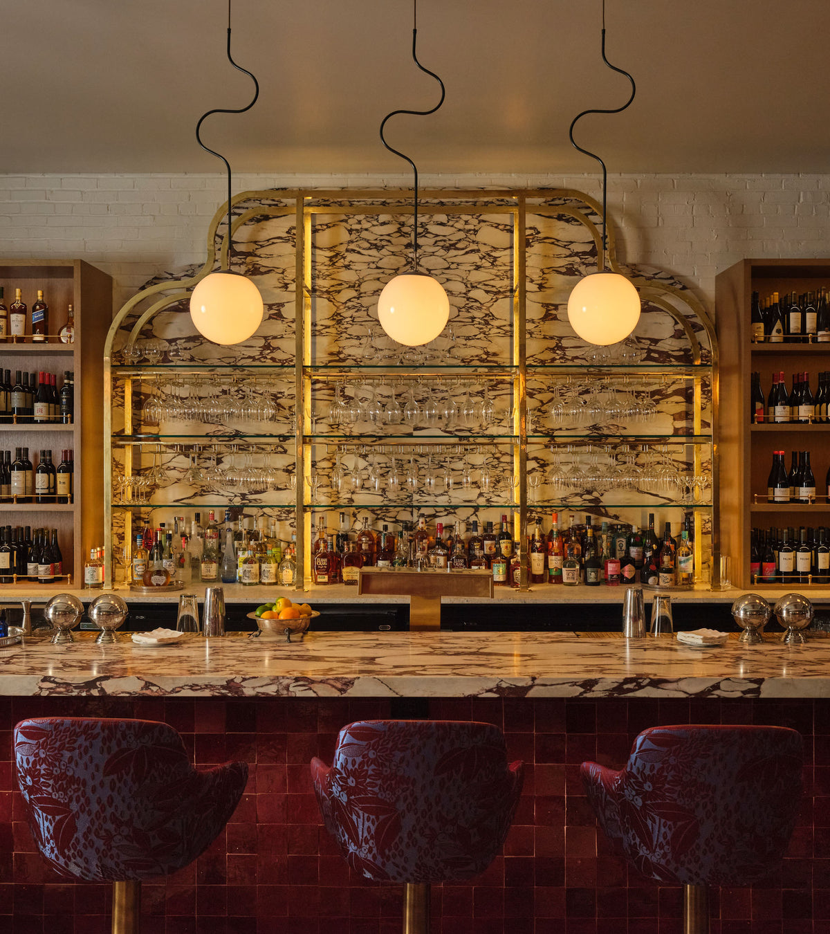 A stylish bar with three red patterned chairs, a marble counter, shelves of bottles and glassware, and globe pendant lights illuminating a gold-accented mirrored wall adorned with vendor-unknown Rouge 4x4 tiles.