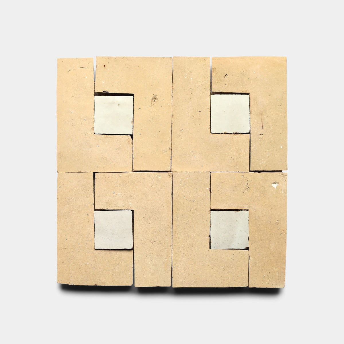 Four Rubric 2, Sample tiles by Zia Tile are arranged in a grid, each beige tile featuring an L-shaped cutout with a white square inlay. The handmade cement tiles create a geometric pattern against a plain light gray background.