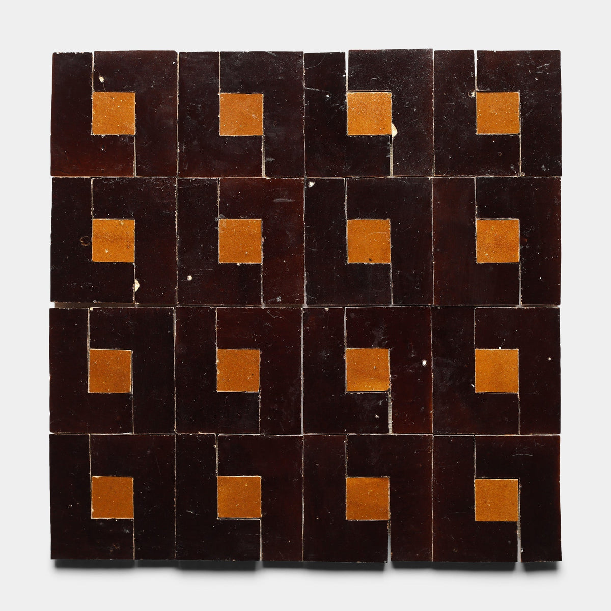 A 4x4 grid of Rubric 5, Sample tiles from Zia Tile features dark brown squares each with an ochre accent in one corner, forming a striking geometric pattern on a white background.