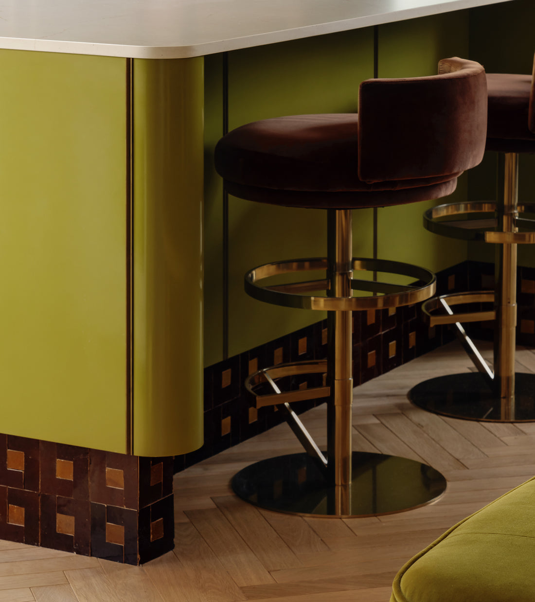 A modern bar area features olive green paneling, a light countertop, brown velvet bar stools with gold bases, and herringbone wood flooring. Zia Tile's Rubric 5 adds Moroccan-inspired geometric brown and gold accents at the counter’s base.