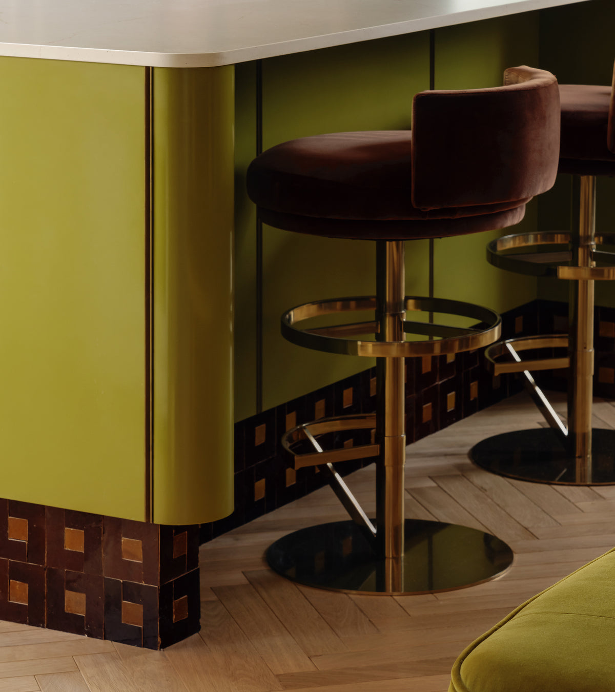 A modern bar area features olive green paneling, a light countertop, brown velvet bar stools with gold bases, and herringbone wood flooring. Zia Tile's Rubric 5 adds Moroccan-inspired geometric brown and gold accents at the counter’s base.