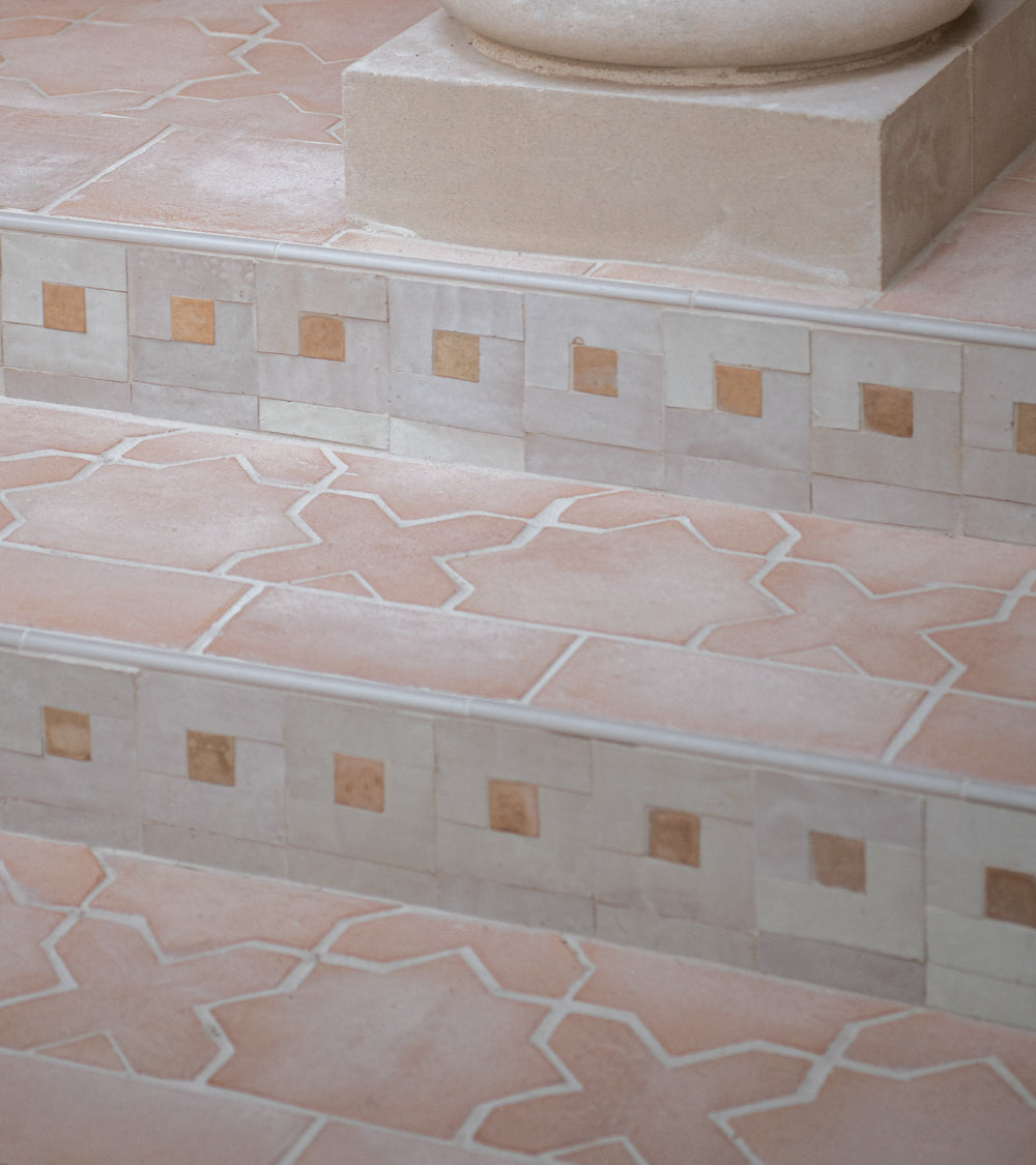 Close-up of tiled steps with Rubric 1 mosaic zellige tiles by Zia Tile, featuring a geometric tan and cream pattern with square accents and a decorative border, leading up to a stone pedestal.