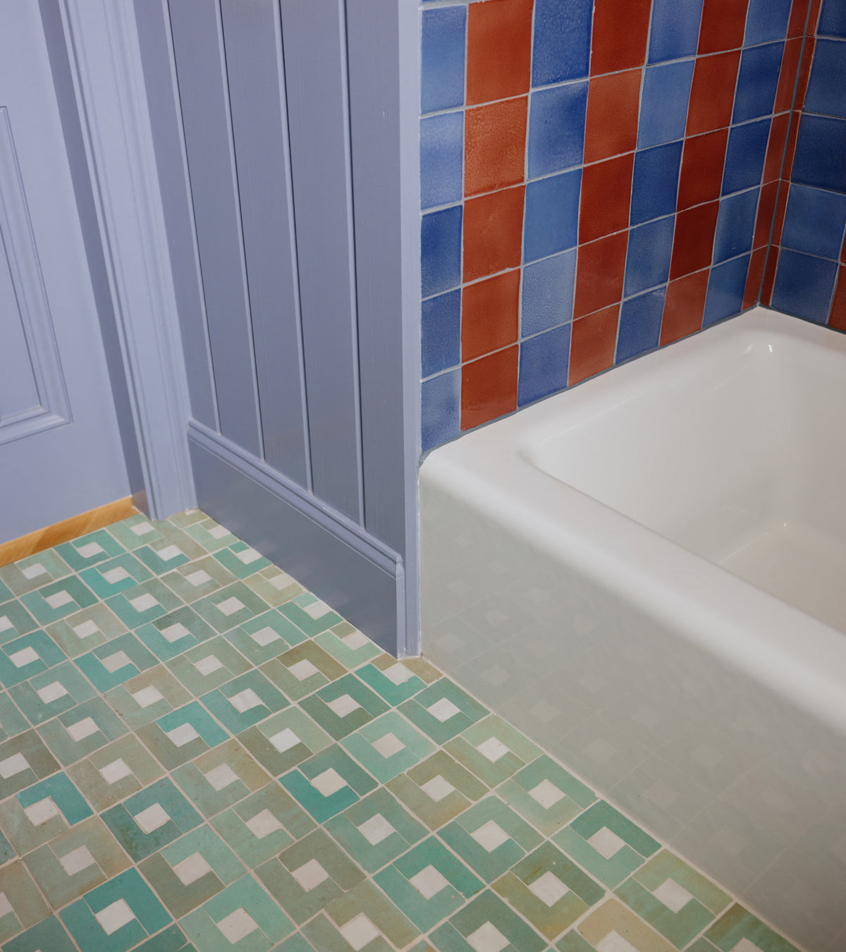 A white bathtub sits beside a wall of blue and rust-colored Rubric 4 tiles by Zia Tile, next to a floor featuring a grid of small light green and white mosaic zellige tiles.