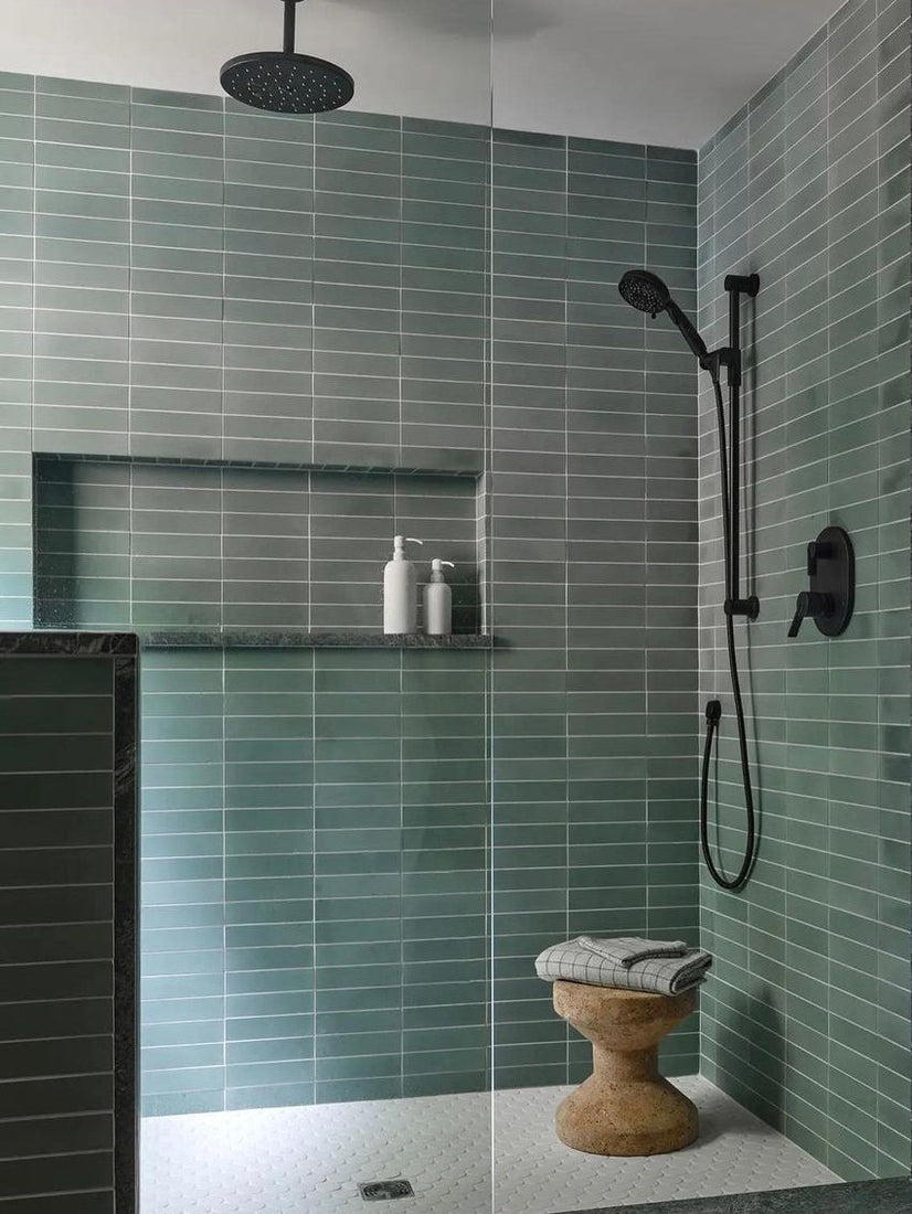 A modern shower features Sage 2x8 tile by vendor-unknown, a black rain showerhead, black handheld shower, built-in shelf with white bottles, a small towel, and a round stool topped with a folded towel.