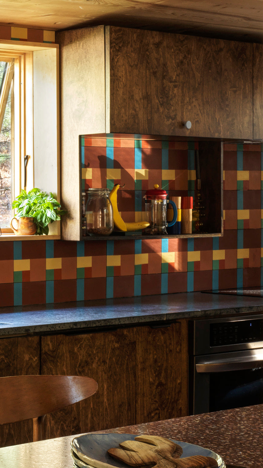 A cozy kitchen with wood cabinets, a colorful geometric tile backsplash in red, blue, yellow, and brown, a window with sunlight, and a small plant on the windowsill. Shelves hold jars, a banana, and a mug.