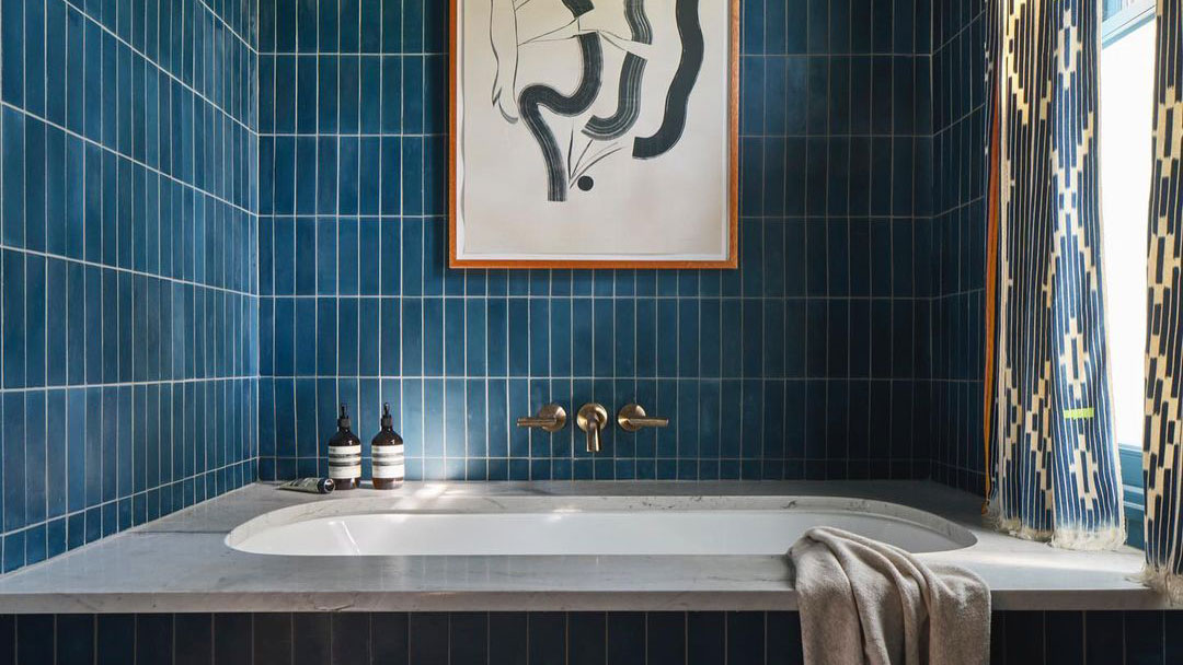 A modern bathroom with blue tiled walls, a white bathtub, two bottles on the tub’s edge, a beige towel draped over the tub, abstract wall art above, and patterned curtains partially visible on the right.