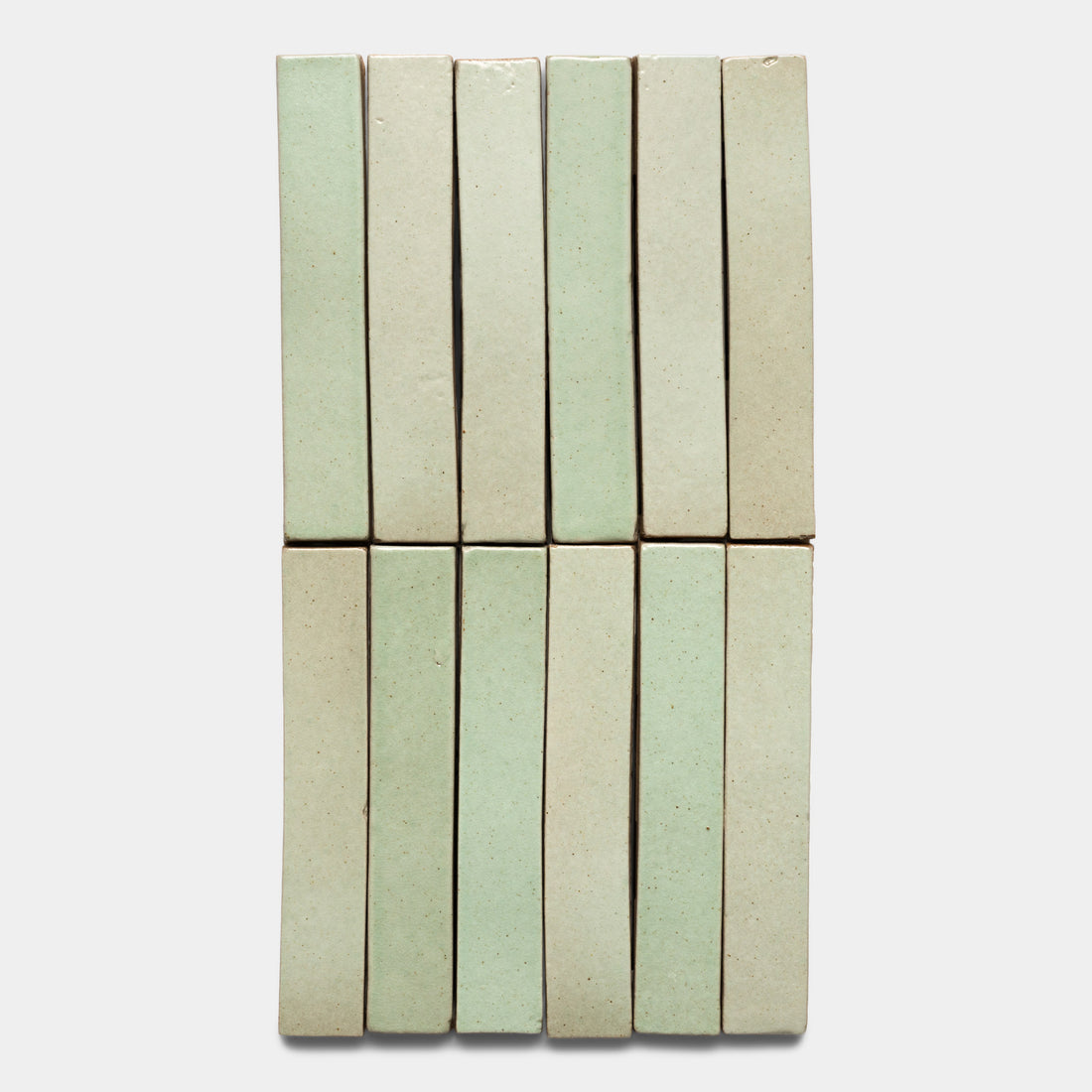 Twelve Sayulita Mini Bar tiles by Zia Tile, arranged vertically in two rows of six, feature a glossy pale green and beige finish against a plain light background.