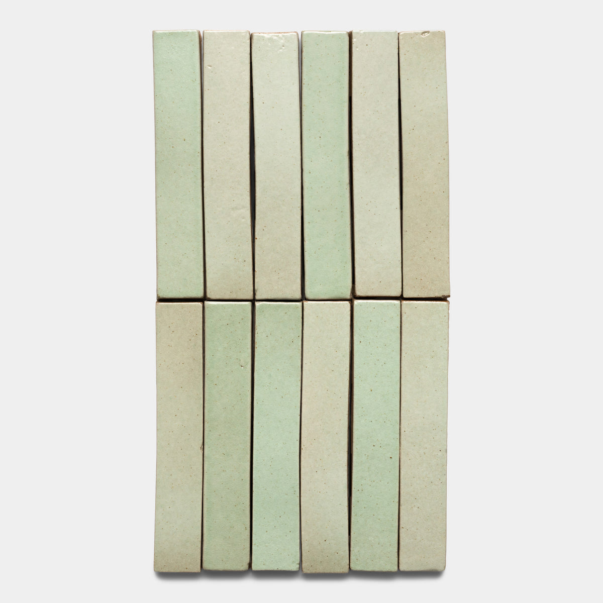 Twelve Sayulita Mini Bar tiles by Zia Tile, arranged vertically in two rows of six, feature a glossy pale green and beige finish against a plain light background.