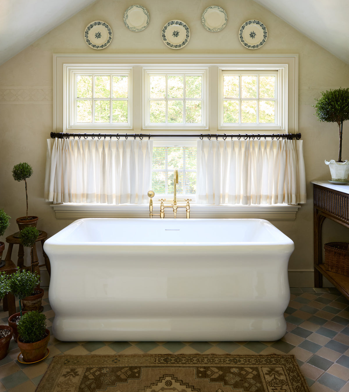 A bright bathroom features a white freestanding tub beneath wide windows with sheer curtains. Sayulita 4x4 Square tiles by Zia Tile and potted topiary plants evoke San Miguel de Allende’s cozy, elegant charm.