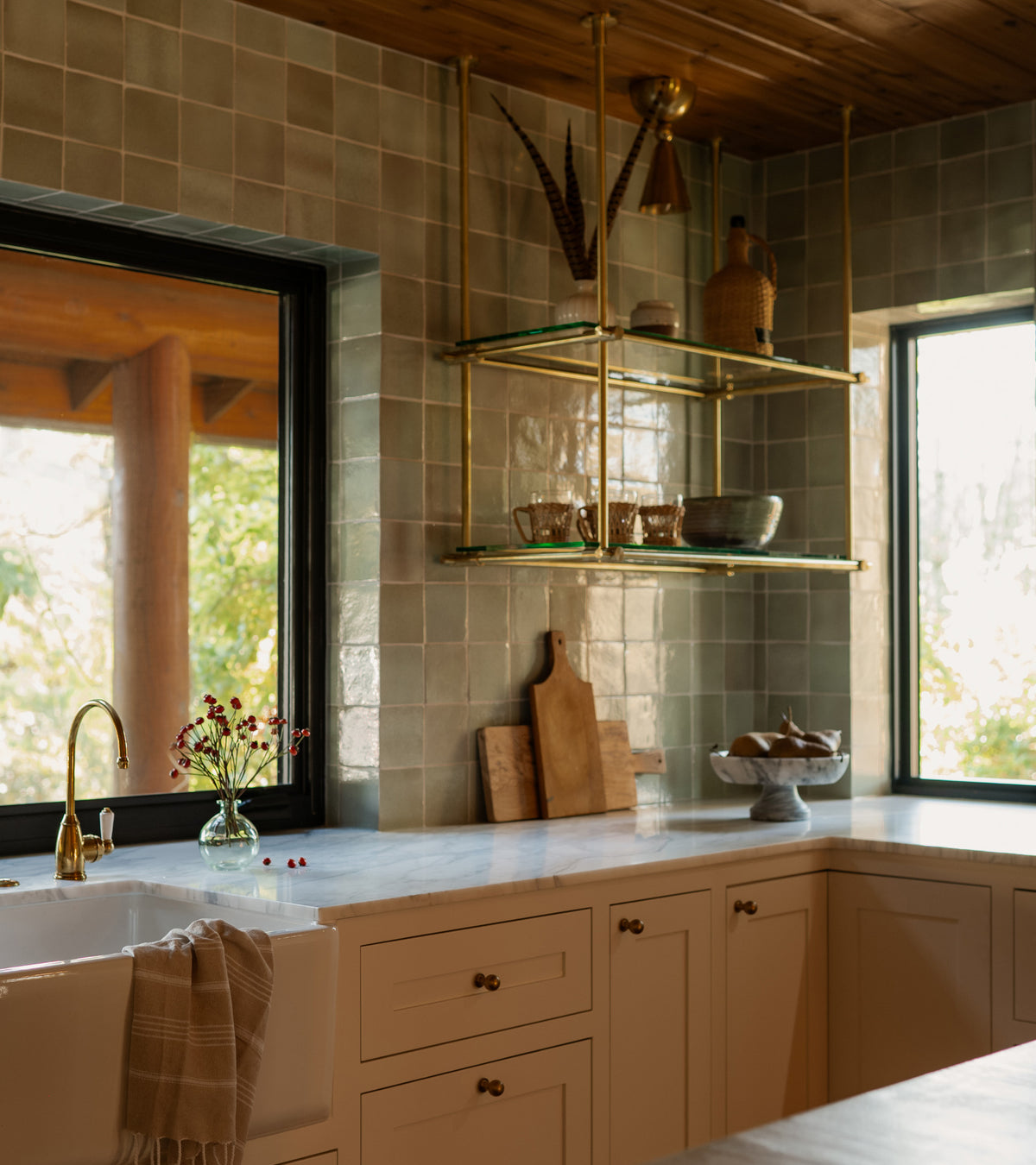 A warm kitchen with cream cabinets, a white farmhouse sink, gold faucet, Zia Tile’s Sayulita 4x4 Square handmade Cotto tile backsplash, open glass shelves with pottery and kitchenware, cutting boards, and large sunlit windows.