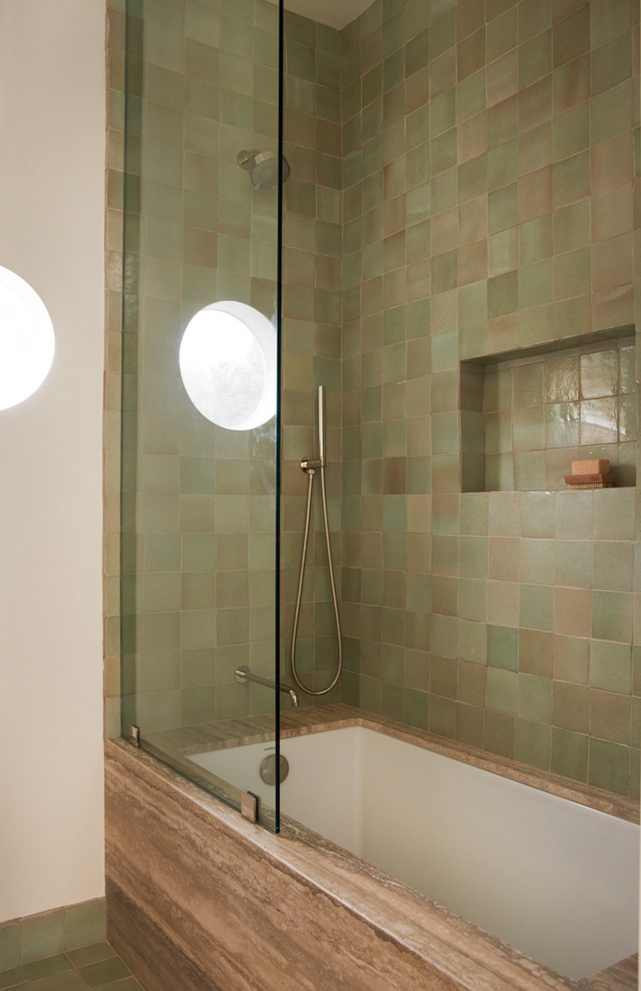 A modern bathroom featuring Zia Tile's Sayulita 4x4 Square green tiles, handmade Cotto accents, a built-in tub with stone edge, glass shower screen, round window, wall-mounted showerhead, and soap on a niche shelf.