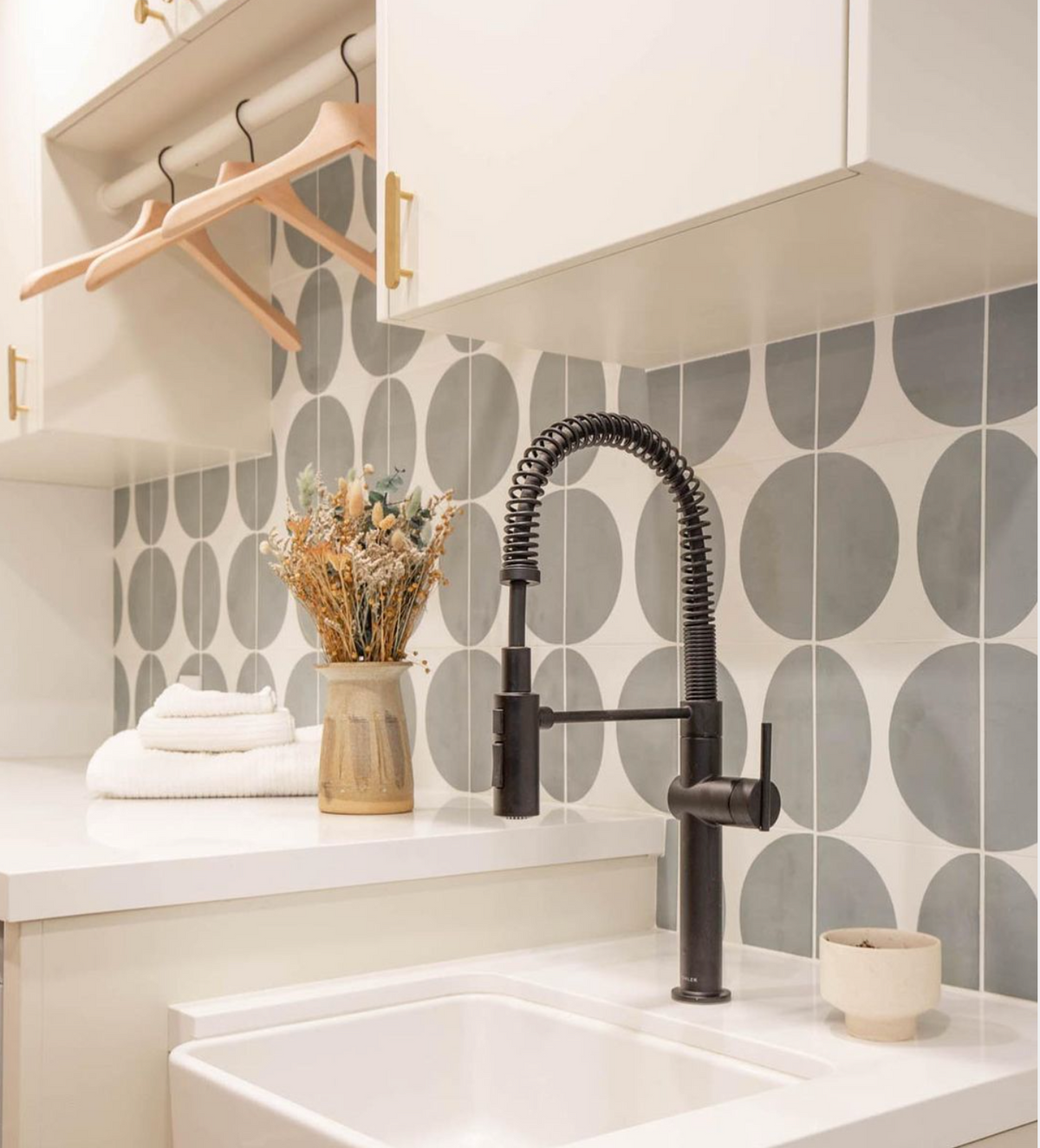 A modern laundry room features a matte black faucet, Pomelo Zeppelin 8x8 tile backsplash by vendor-unknown, stacked white towels, dried flowers in a vase, and a wooden hanging rack under white cabinets.