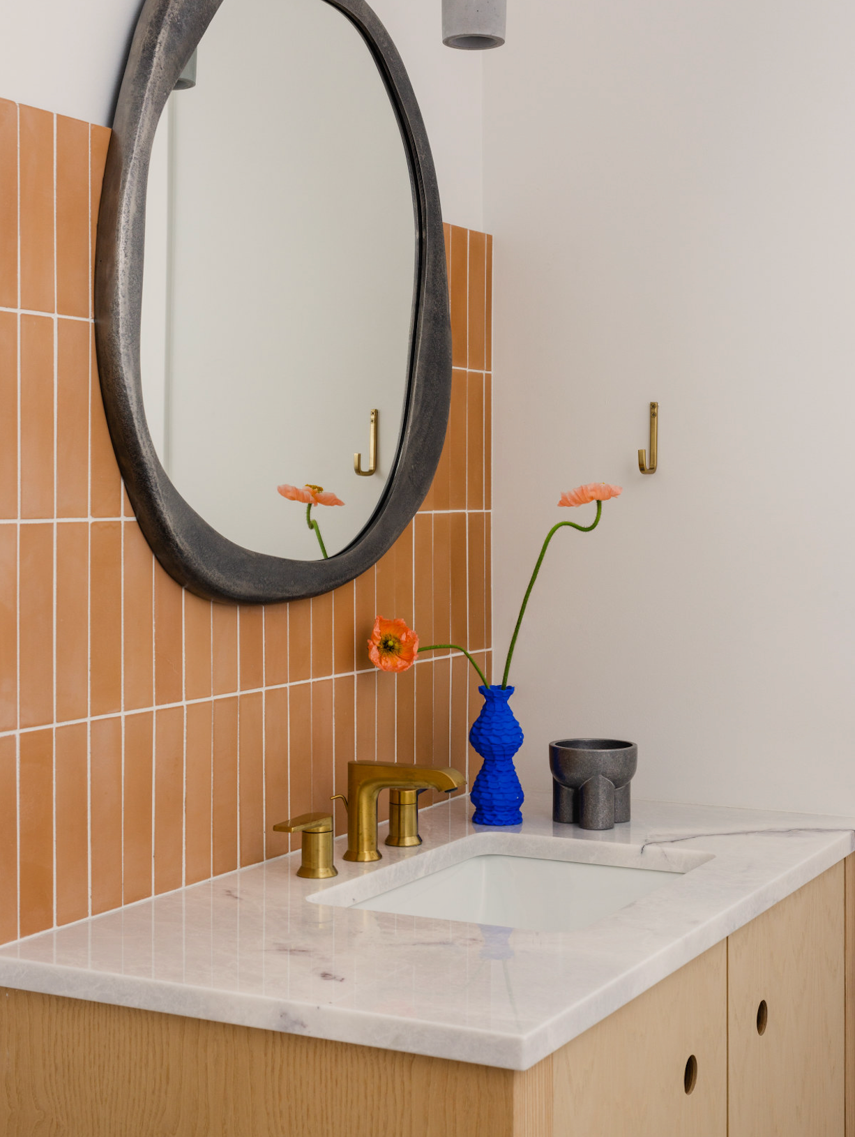 A modern bathroom features a round mirror above a marble sink counter, brass fixtures, and a Terra Cotta 2x8 tile backsplash. A blue vase with orange flowers brings a vibrant accent.