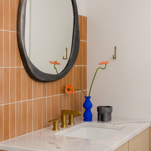 A modern bathroom features a round mirror above a marble sink counter, brass fixtures, and a Terra Cotta 2x8 tile backsplash. A blue vase with orange flowers brings a vibrant accent.