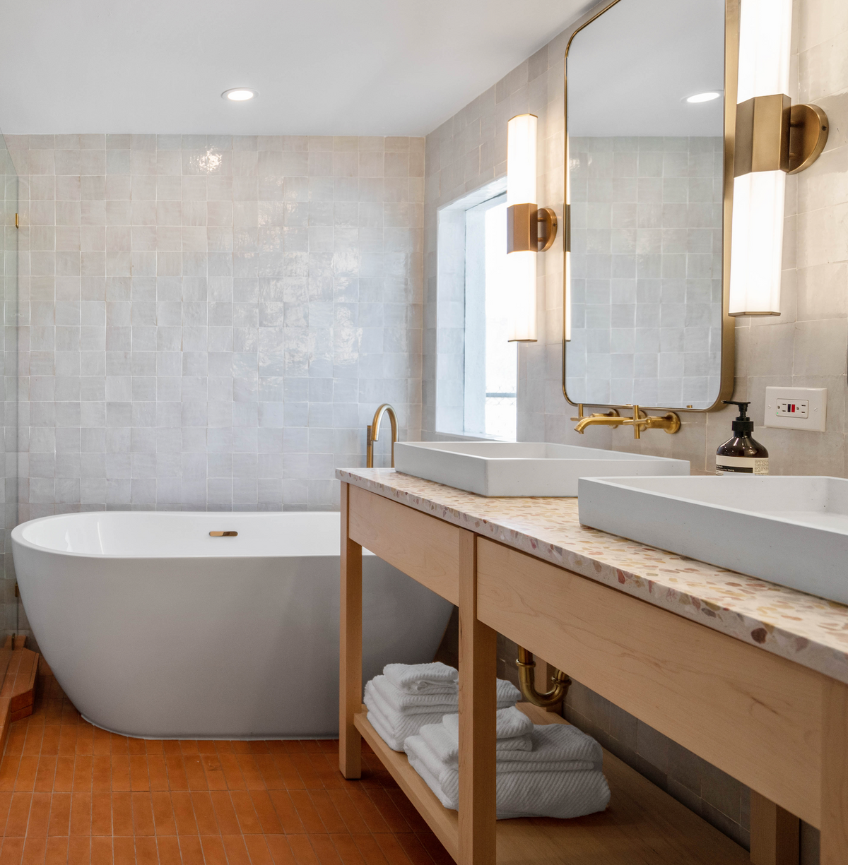 Modern bathroom with a white freestanding tub, double sinks on a light wood vanity, gold fixtures, and folded white towels on a lower shelf. Terra Cotta 2x8 tiles add a sleek touch to the walls beside the tall mirror.