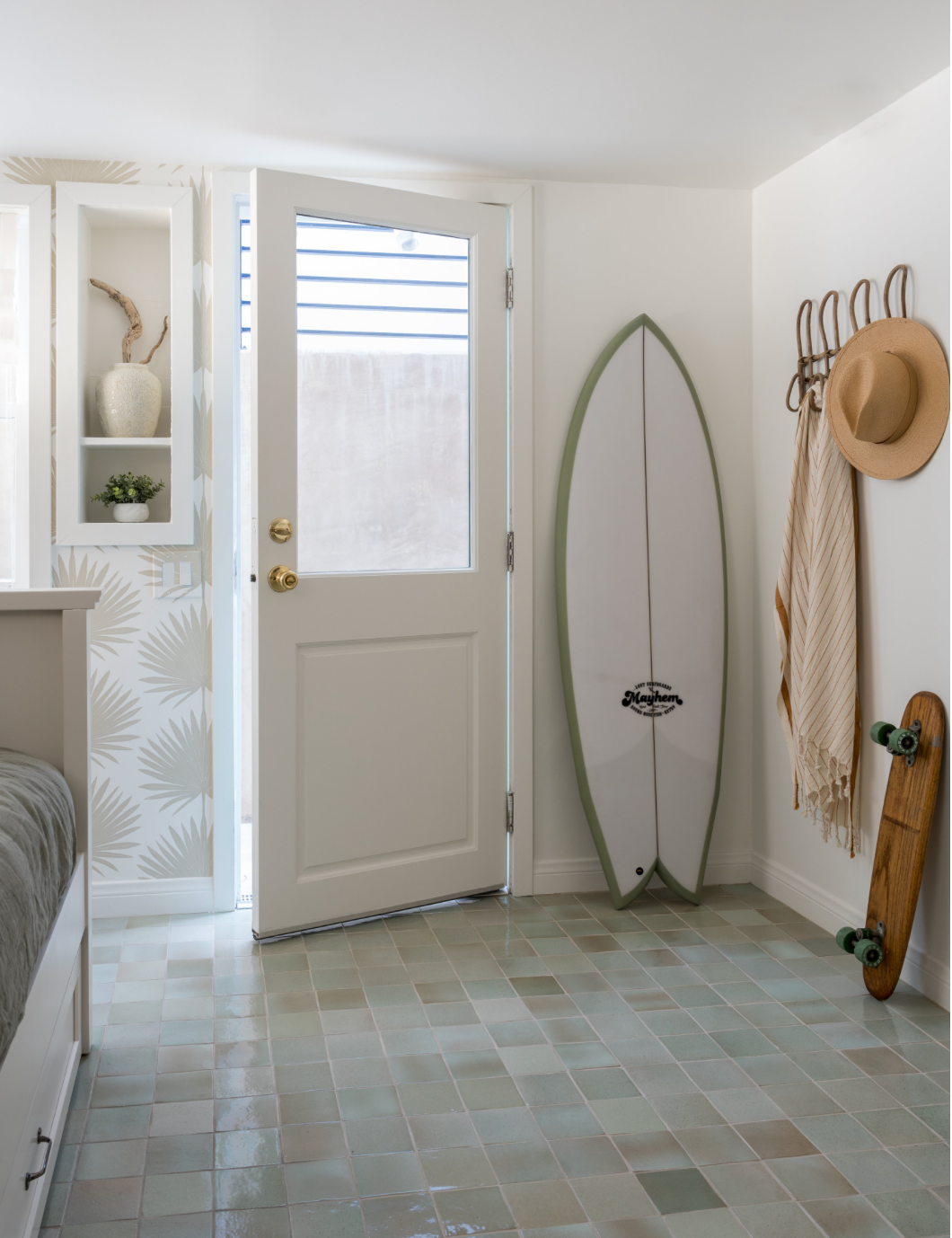 A bright, coastal-themed entryway features a white door, surfboard, skateboard, hooks with a hat and striped towels, shelving with decor, and a pastel Sayulita 4x4 Square floor tile by Zia Tile.