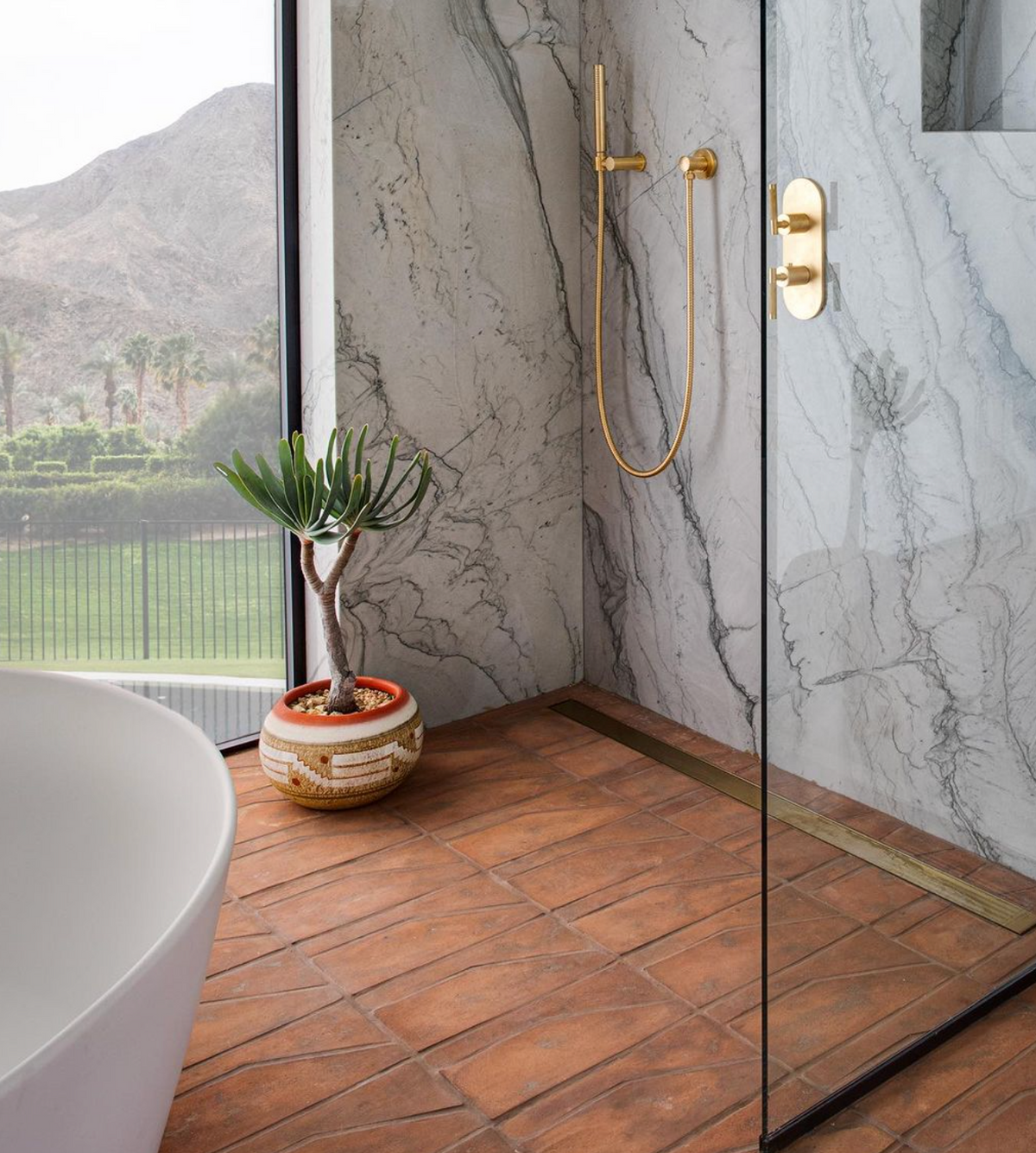 A modern bathroom featuring Zocalo + Fired Earth terracotta tile by Zia Tile, a marble shower, gold fixtures, white bathtub, potted succulent, and a large window with mountain views.